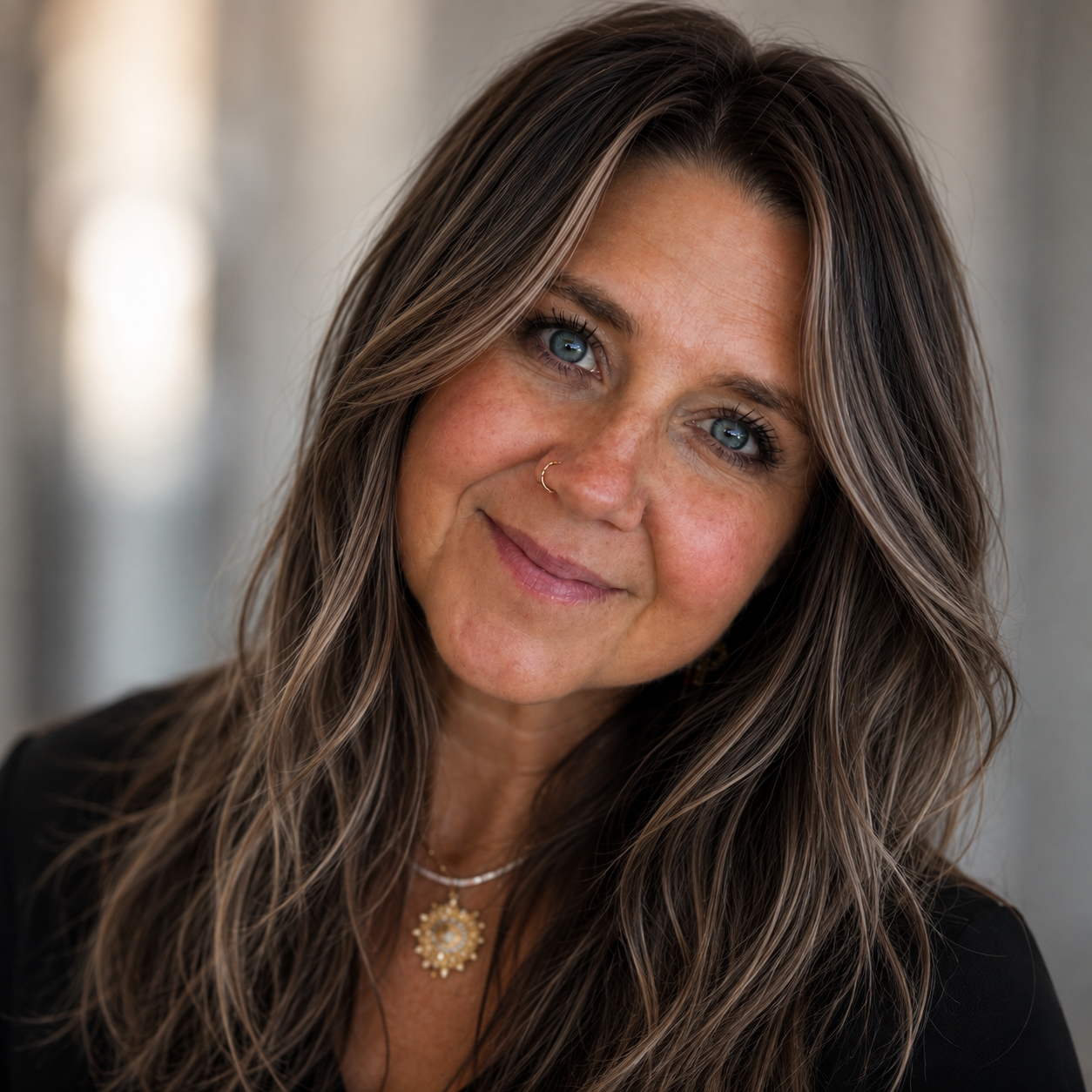 Close-up portrait of a woman with long, wavy brown hair and blue eyes, smiling gently, wearing a gold necklace with a pendant, a nose ring, and a black top, with a blurred neutral background.