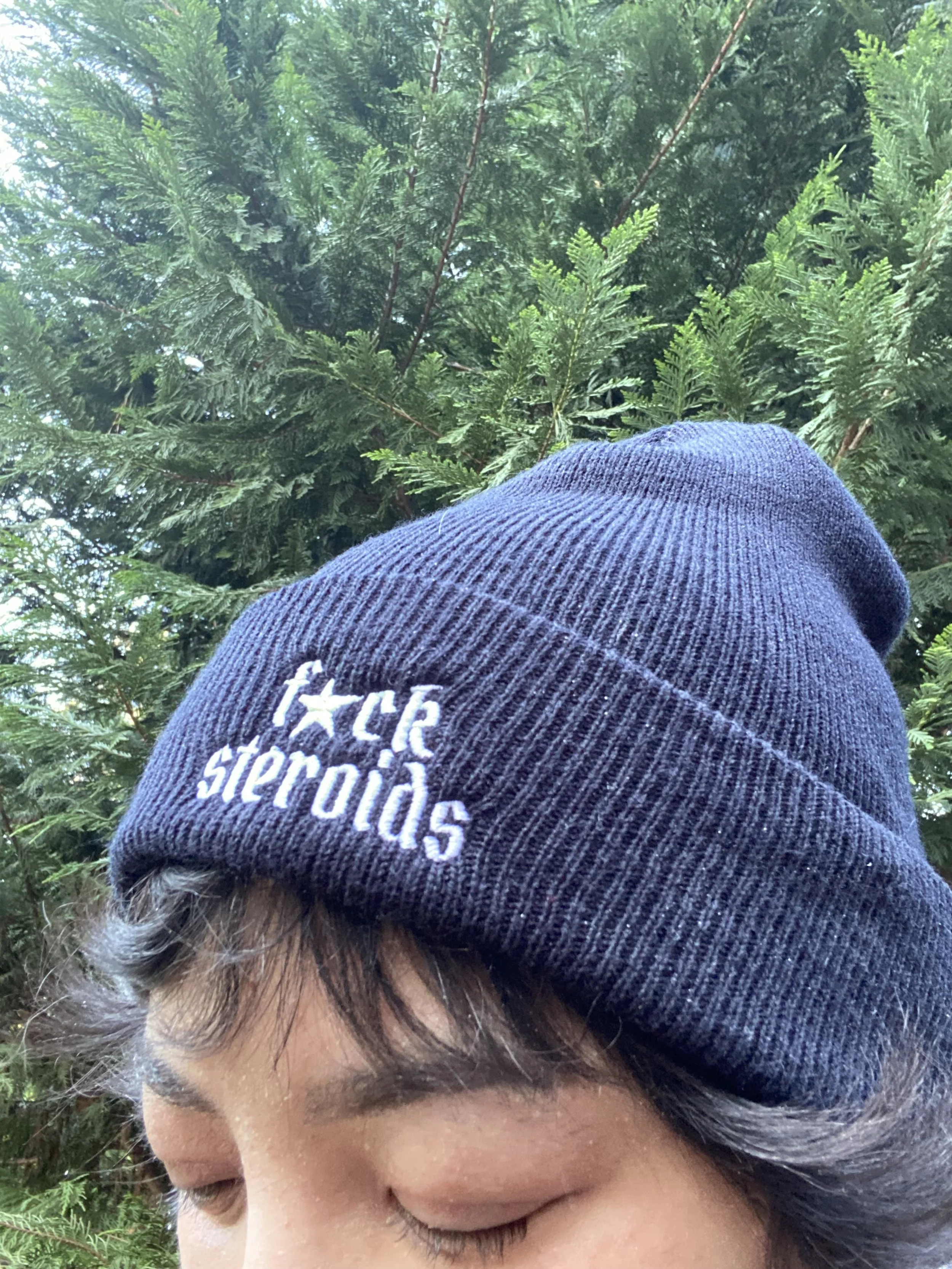 A woman shopping for apples in a grocery store, wearing a black T-shirt that advocates for and explains what topical steroid withdrawal is with white and pink text on the back. She is reaching for apples in a wooden display.
