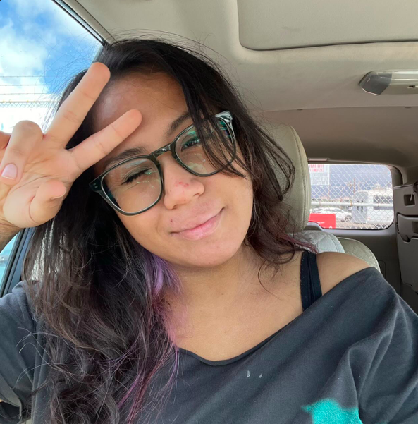 Young woman with glasses making a peace sign inside a vehicle, with a purple streak in her hair and a slight smile.