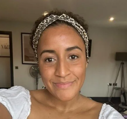 A woman with curly hair wearing a leopard print headband and a white top, smiling in a room with framed pictures and a lamp in the background.