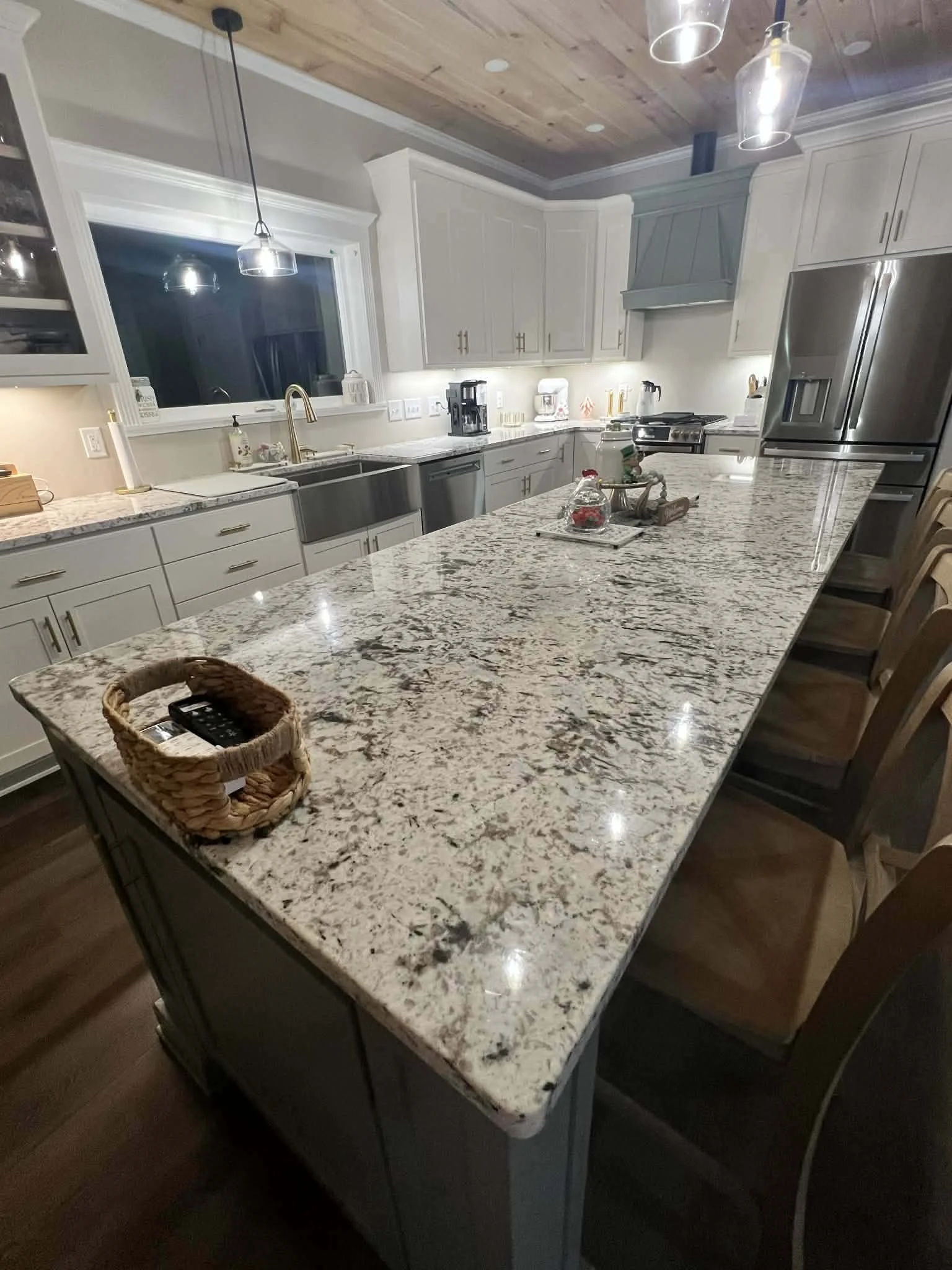 A modern kitchen with a large granite island counter, white cabinets, stainless steel appliances, and pendant lights hanging from a wooden ceiling.