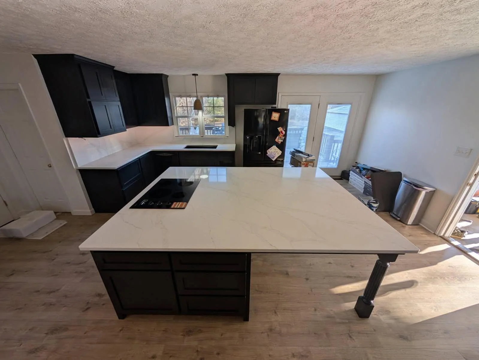 A modern kitchen with black cabinets, a large white marble island, a black stove built into the island, a black refrigerator, and hardwood floors. There are some boxes and a stack of magazines or books near the sliding glass door with a deck outside.