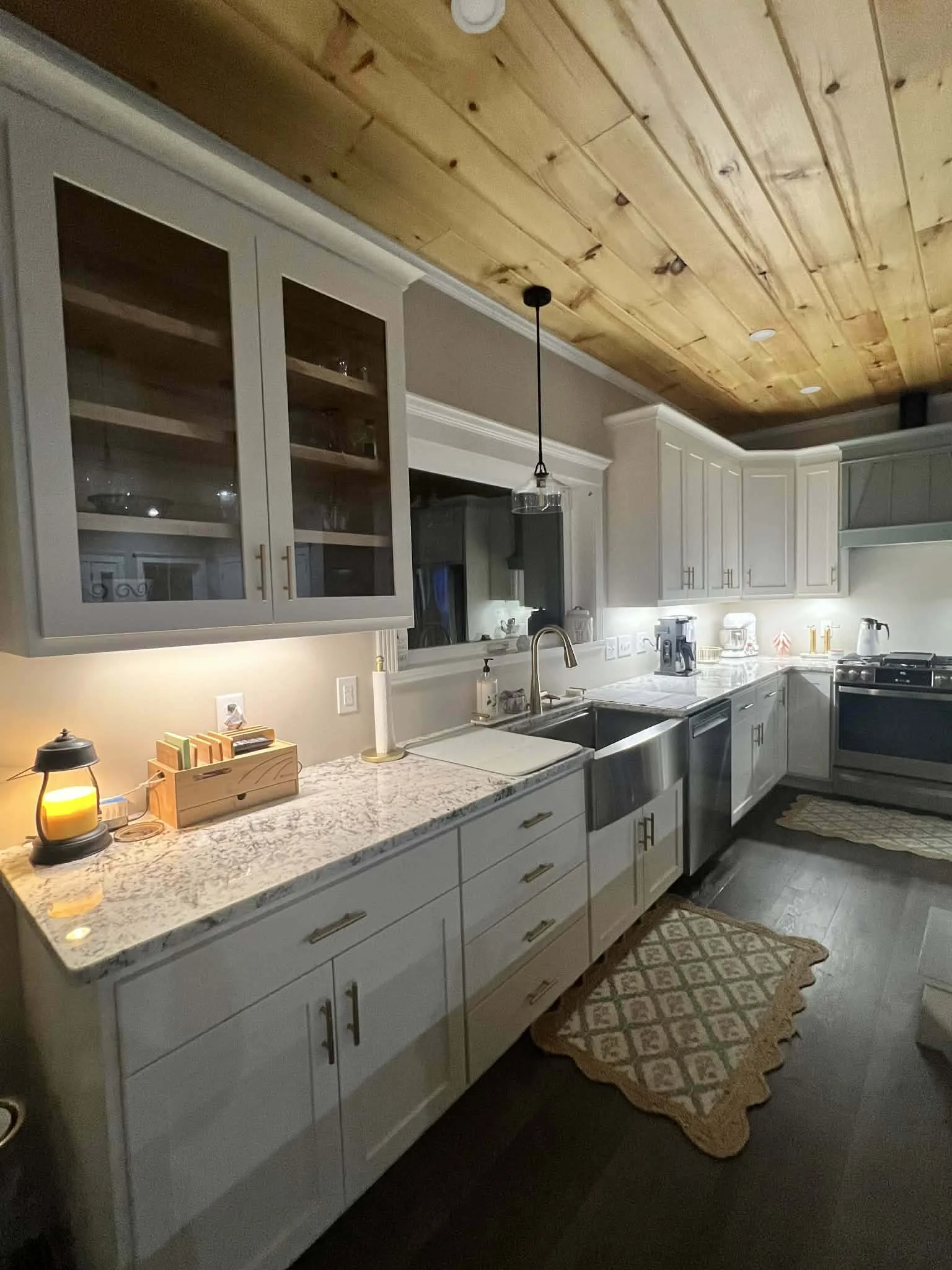 A modern kitchen with white cabinets, granite countertops, and a wooden ceiling. The kitchen includes a double sink, a stove, and various small appliances. There are two small area rugs on the dark hardwood floor and a pendant light hanging over the 