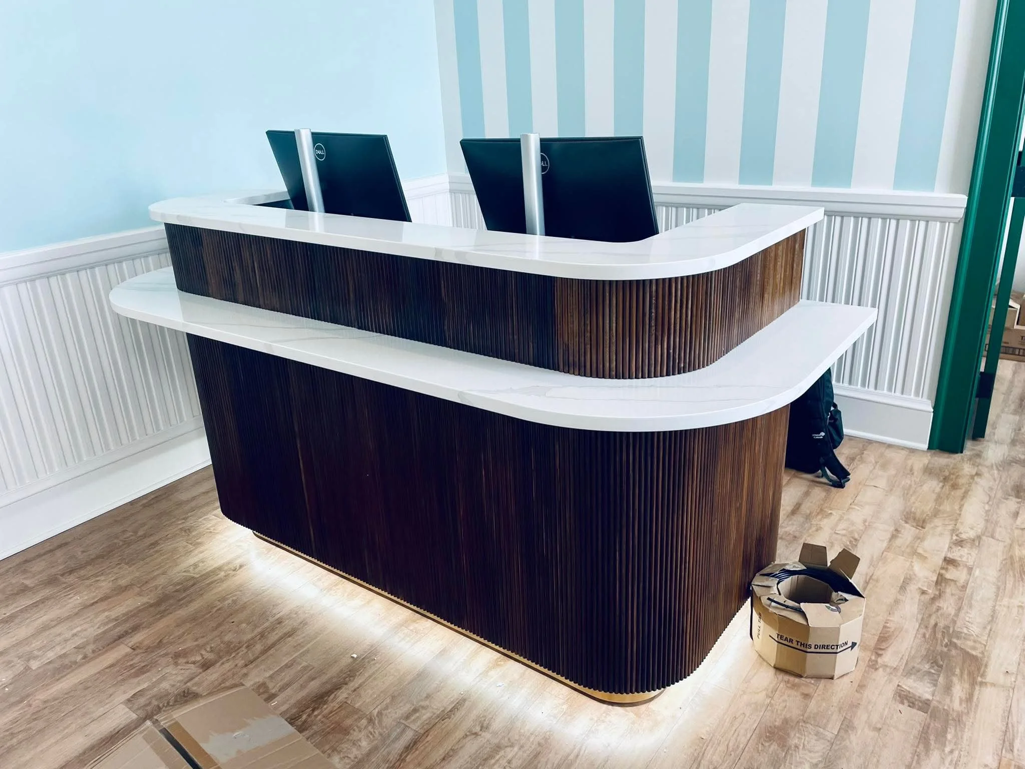 Modern reception desk with two computer monitors and wood paneling, located in a well-lit room with light blue and white striped walls, wooden floor, and a cardboard box on the floor.