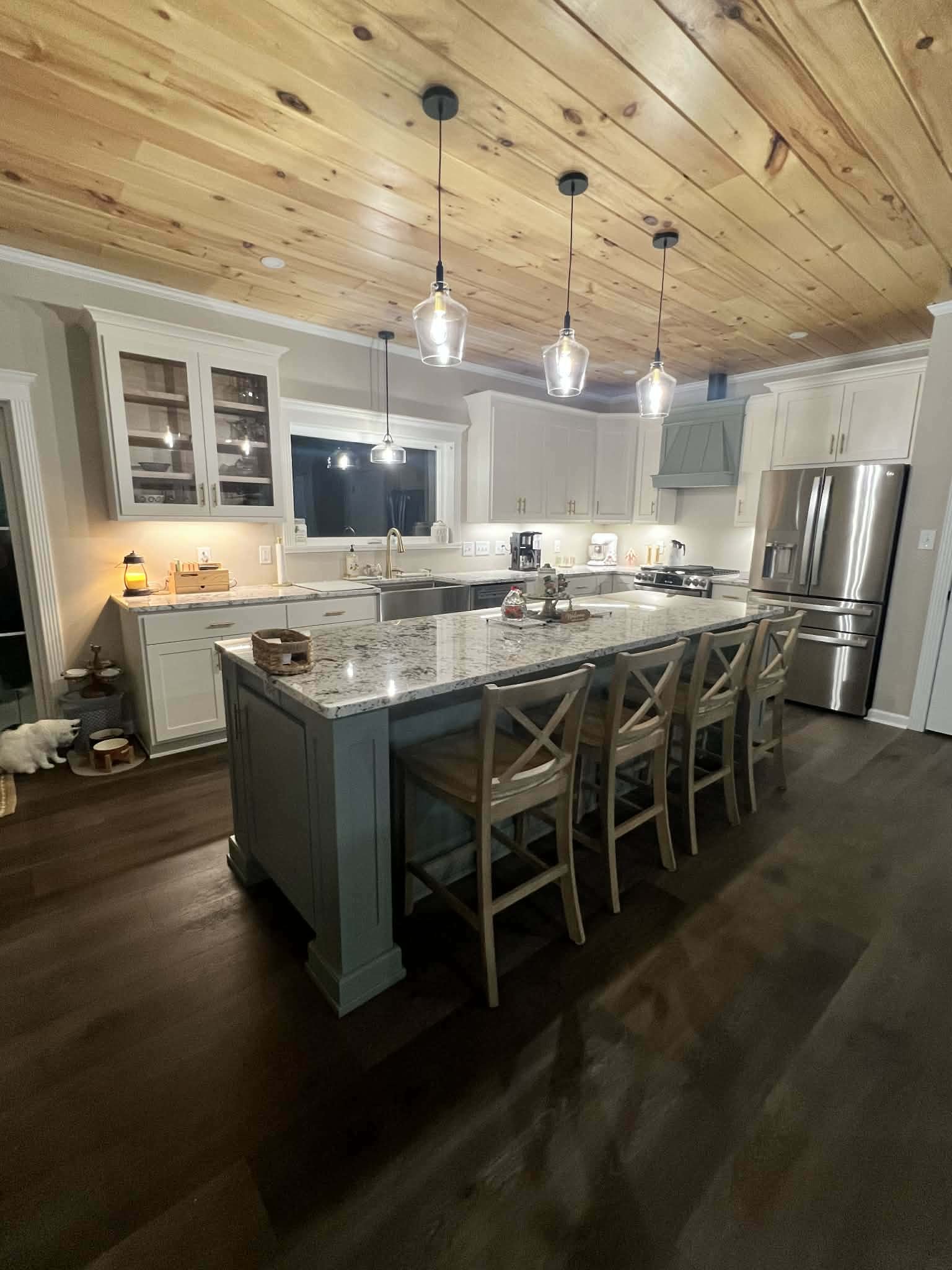 A modern kitchen with a large island topped with granite, white cabinets, stainless steel refrigerator, wooden ceiling, and pendant lights hanging above the island.