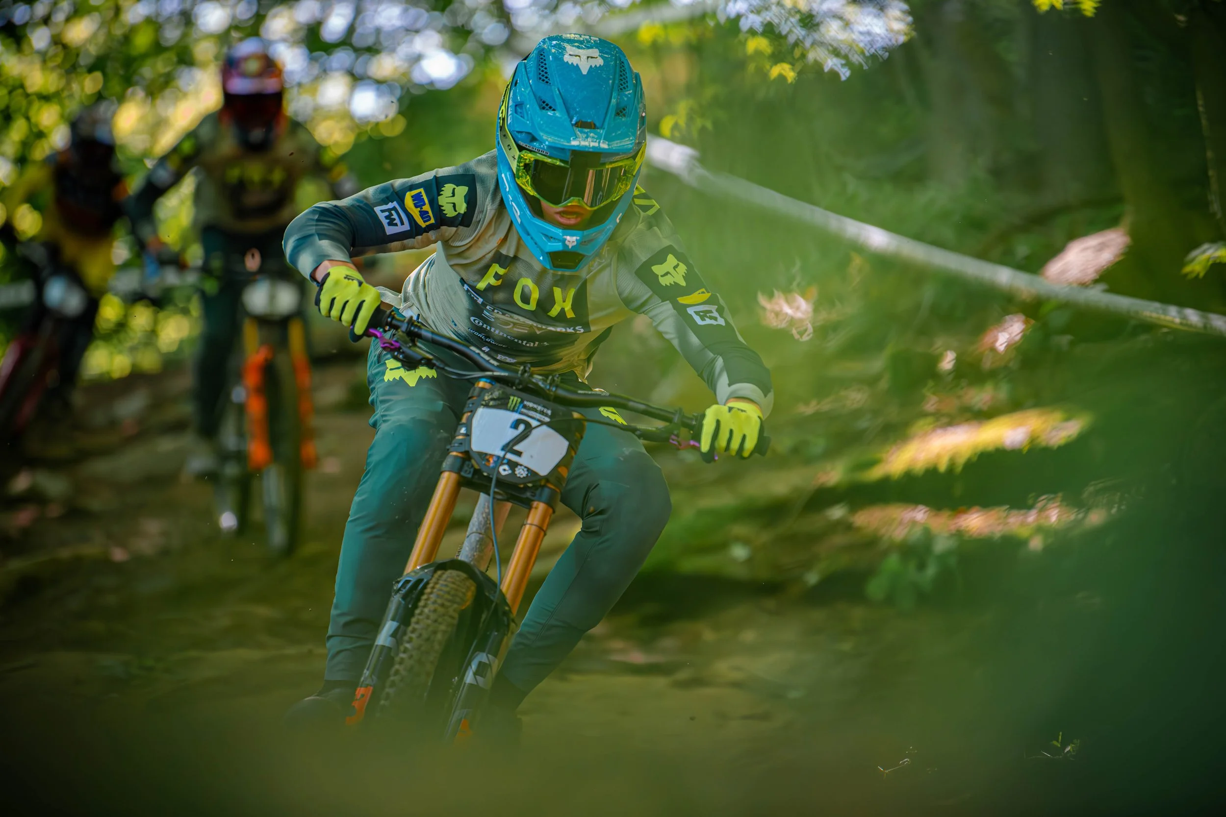 A mountain biker in a blue helmet and yellow gloves racing down a forest trail, with two other mountain bikers following behind.