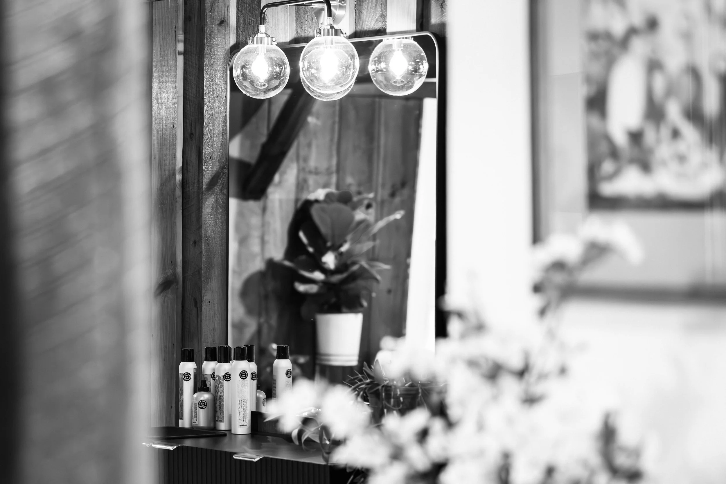 Black and white photo of a wall with light bulbs and a mirror, showing a person with a cap and a tattoo on the arm, standing near a flower pot. Hair care bottles are placed on a table in the foreground.