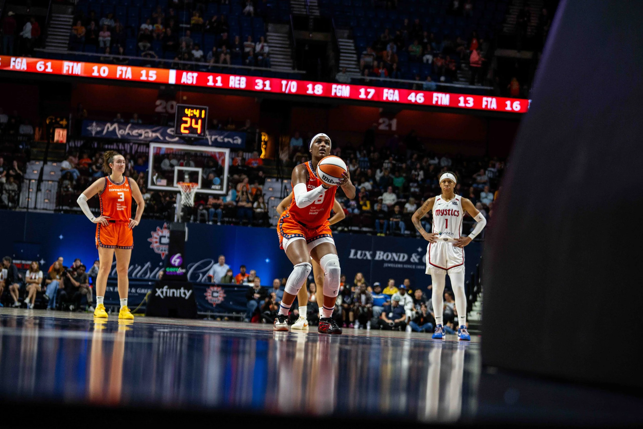 A female basketball player in red uniform is preparing to shoot a free throw on the court, with two other players in white and orange uniforms standing nearby, while spectators watch from the stands.