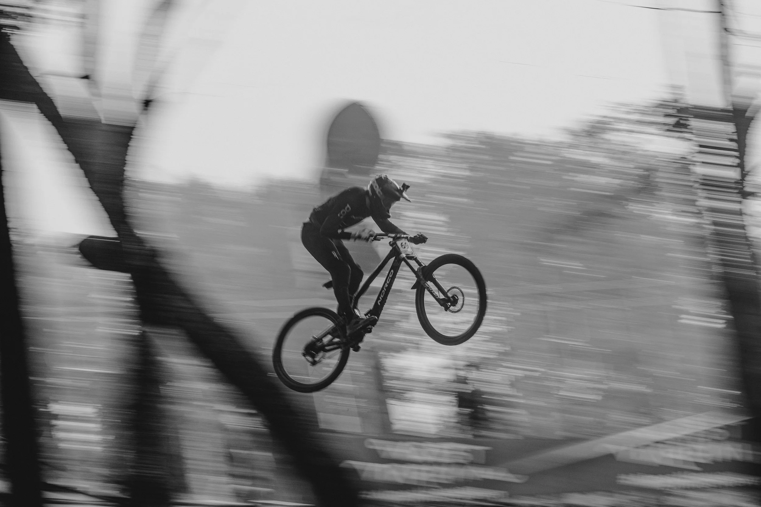 A person riding a mountain bike in mid-air during a jump at a dirt track or BMX course, with the background blurred indicating motion.