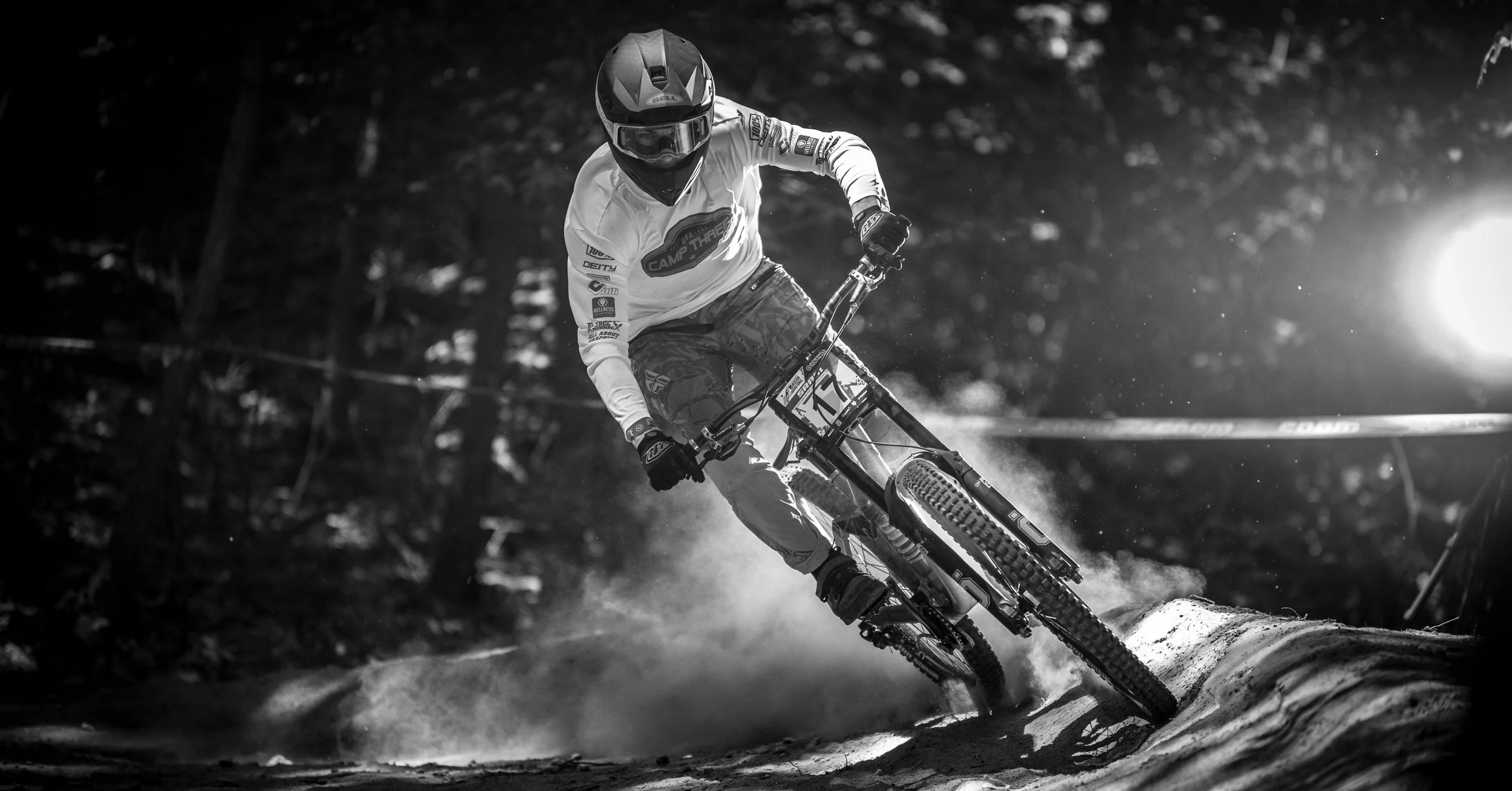A mountain biker wearing a helmet and gloves rides downhill on a dirt trail, kicking up dust, in a forested area at sunset.