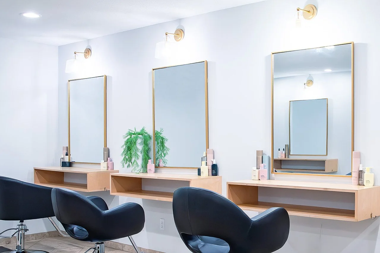 Empty hair salon with three mirrors, black salon chairs, wooden shelves, bottles of hair products, and a green plant