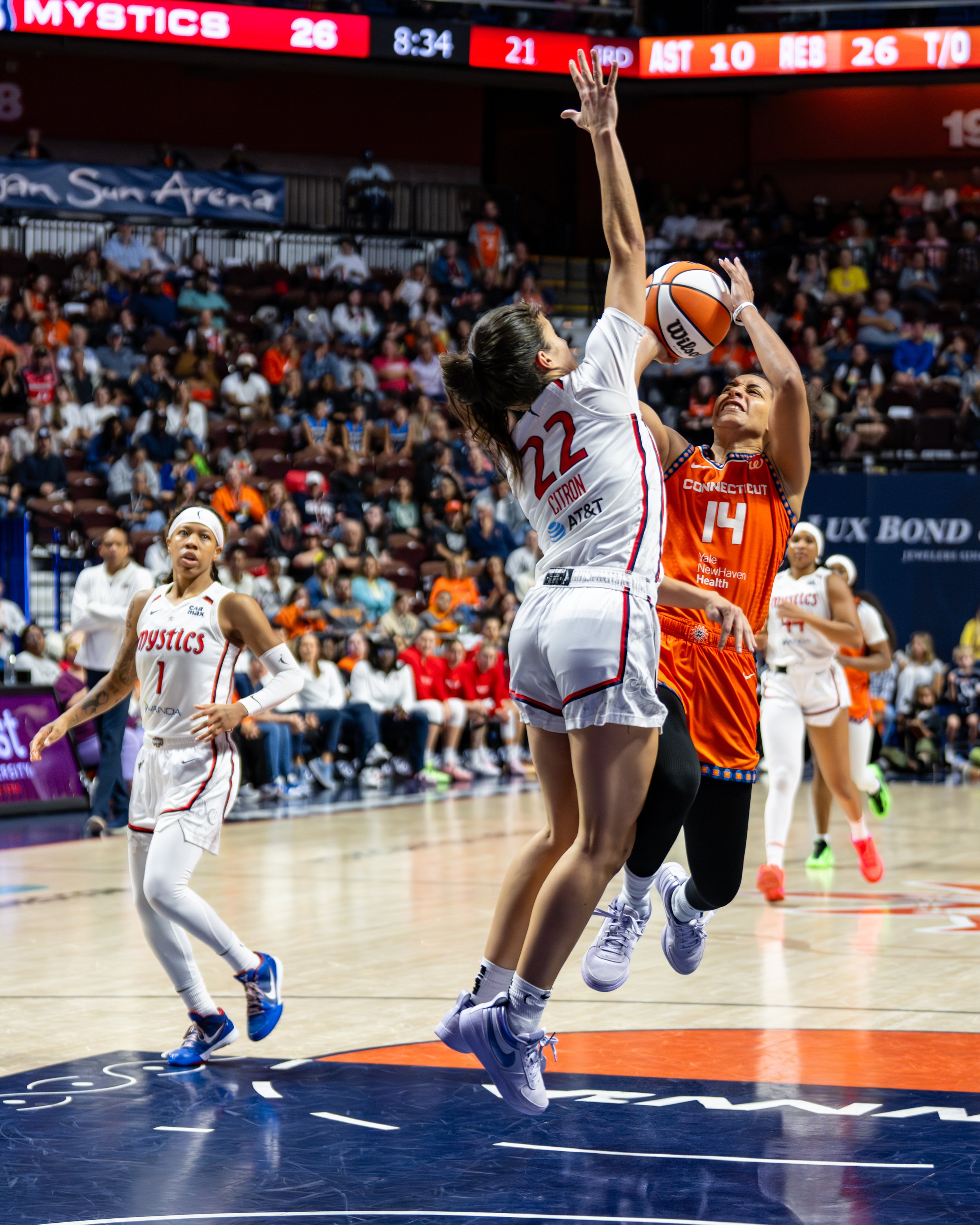 Two female basketball players compete for the ball near the basket, with a crowd of spectators in the background on a basketball court.