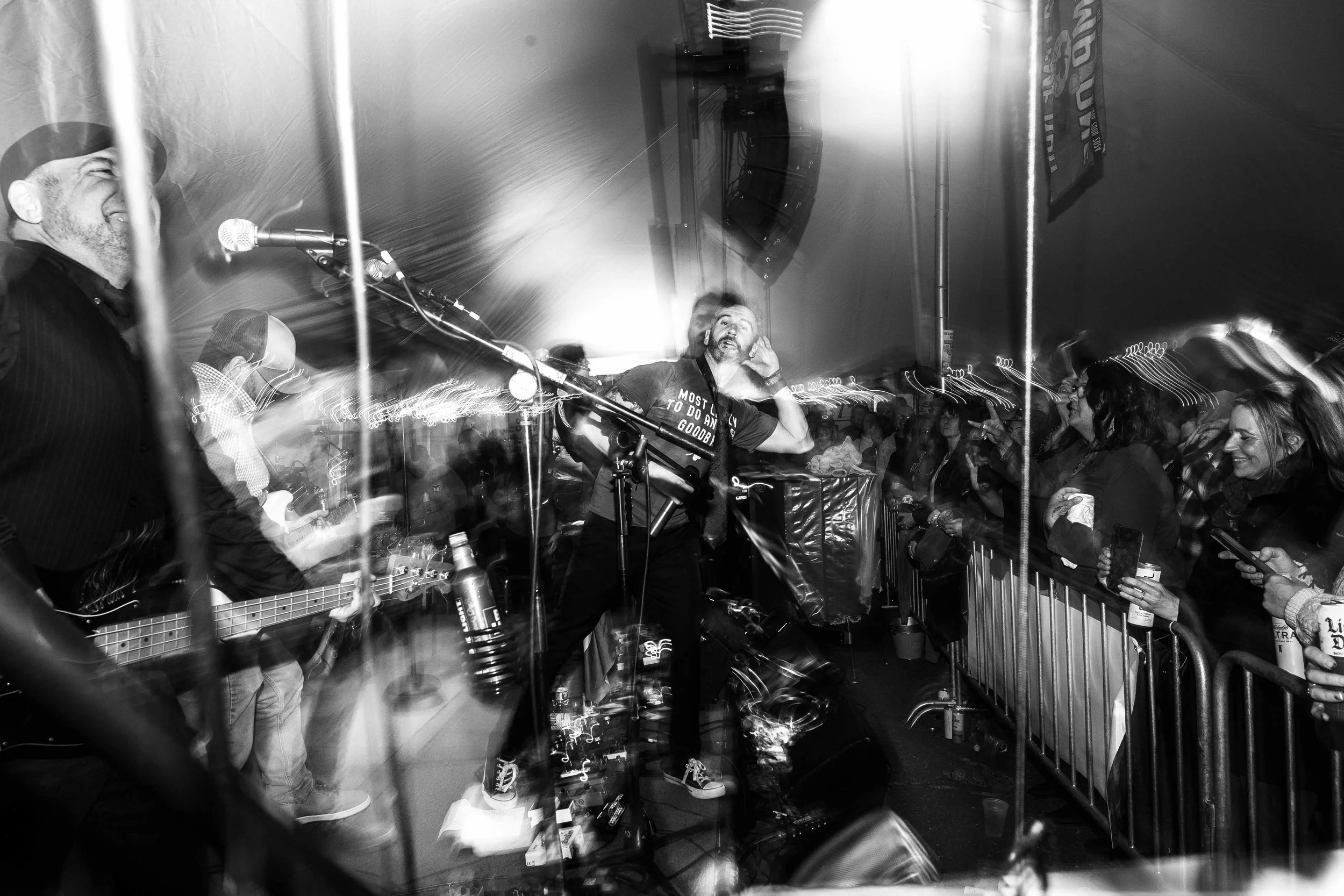 Black and white photo of a band performing on stage at an indoor concert, with audience members standing behind a barrier, some holding drinks and taking photos.