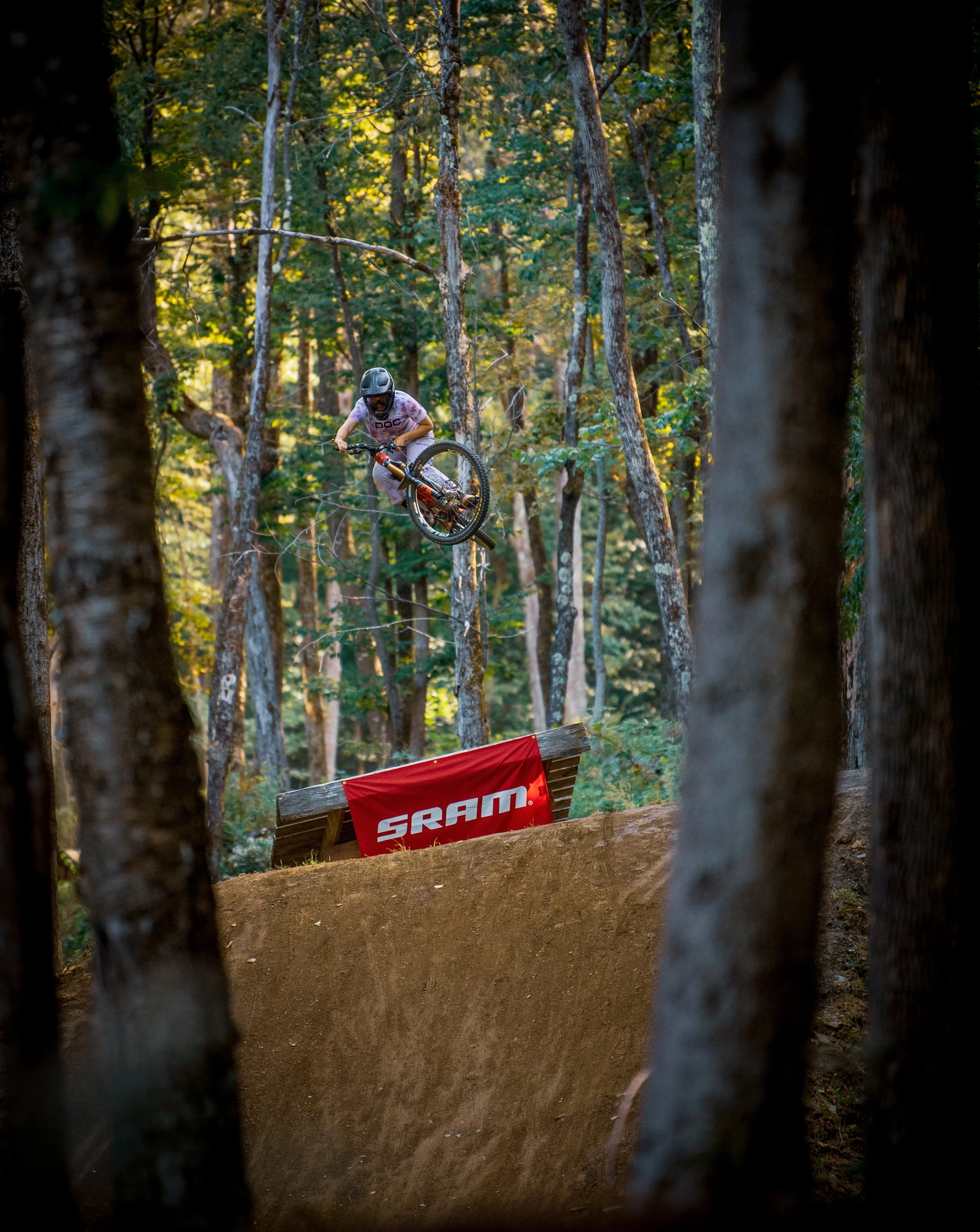 A person performing a jump on a mountain bike in a forested trail with trees and a red SRAM sign in the background.