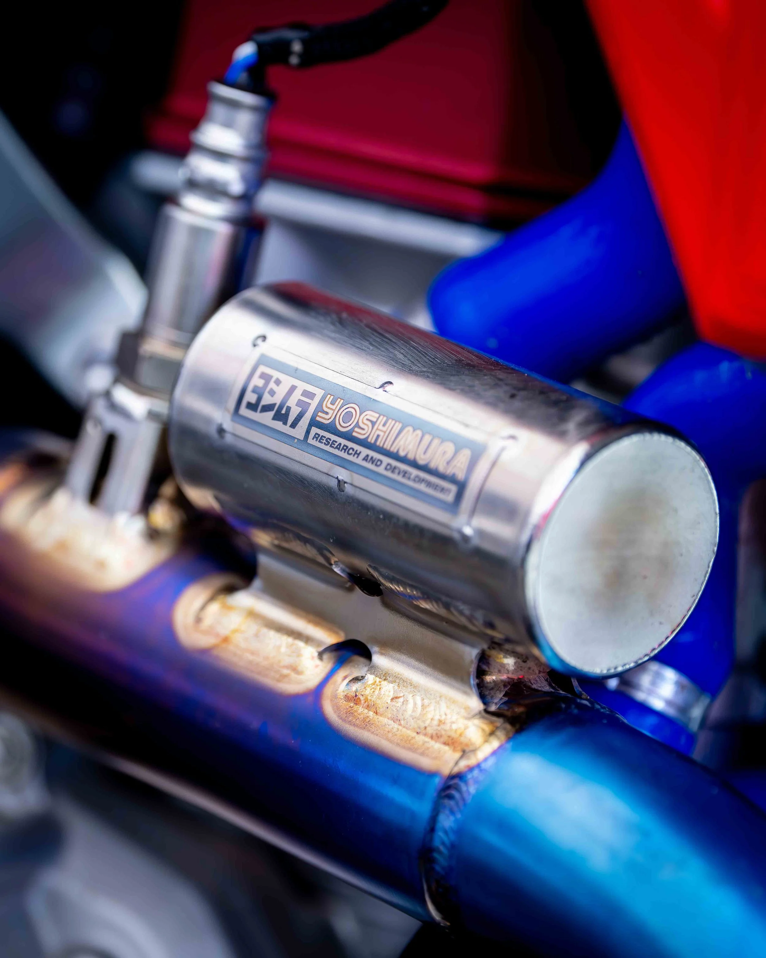 Close-up of a Yoshimura motorcycle exhaust pipe with welded joints, showing a shiny metal surface, attached to blue and red components.