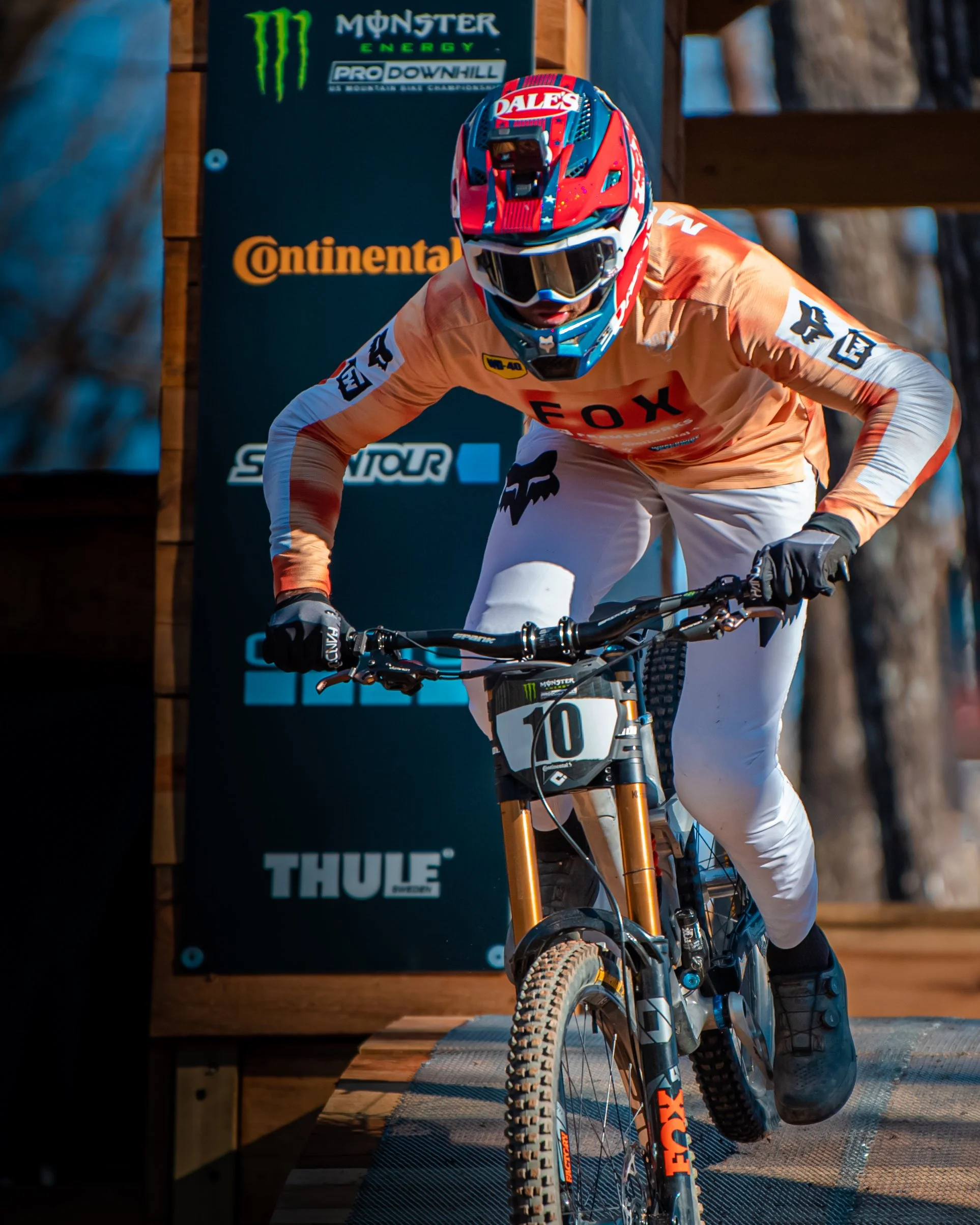 Mountain biker wearing an orange and white FOX jersey and a red, blue, and white helmet riding downhill on a wooden trail with sponsor banners in the background.