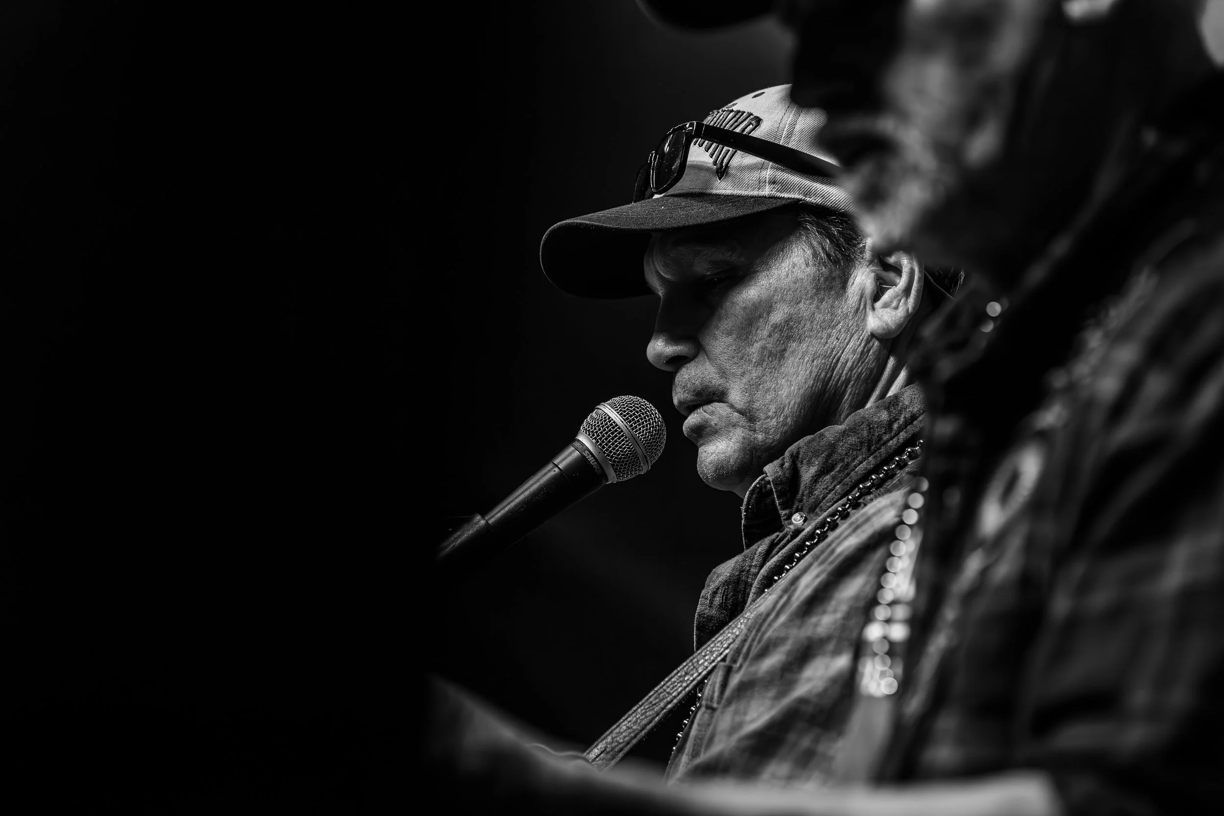 Black and white photo of a man singing into a microphone, wearing a baseball cap and glasses on top, with another person partially visible in the foreground.