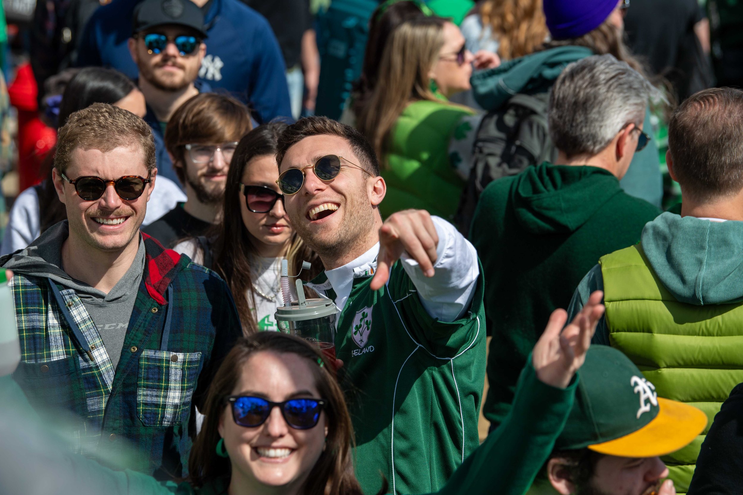 Crowd of people, mostly young adults, gathered outdoors at a sports event, wearing sunglasses and casual clothing, some cheering and smiling.