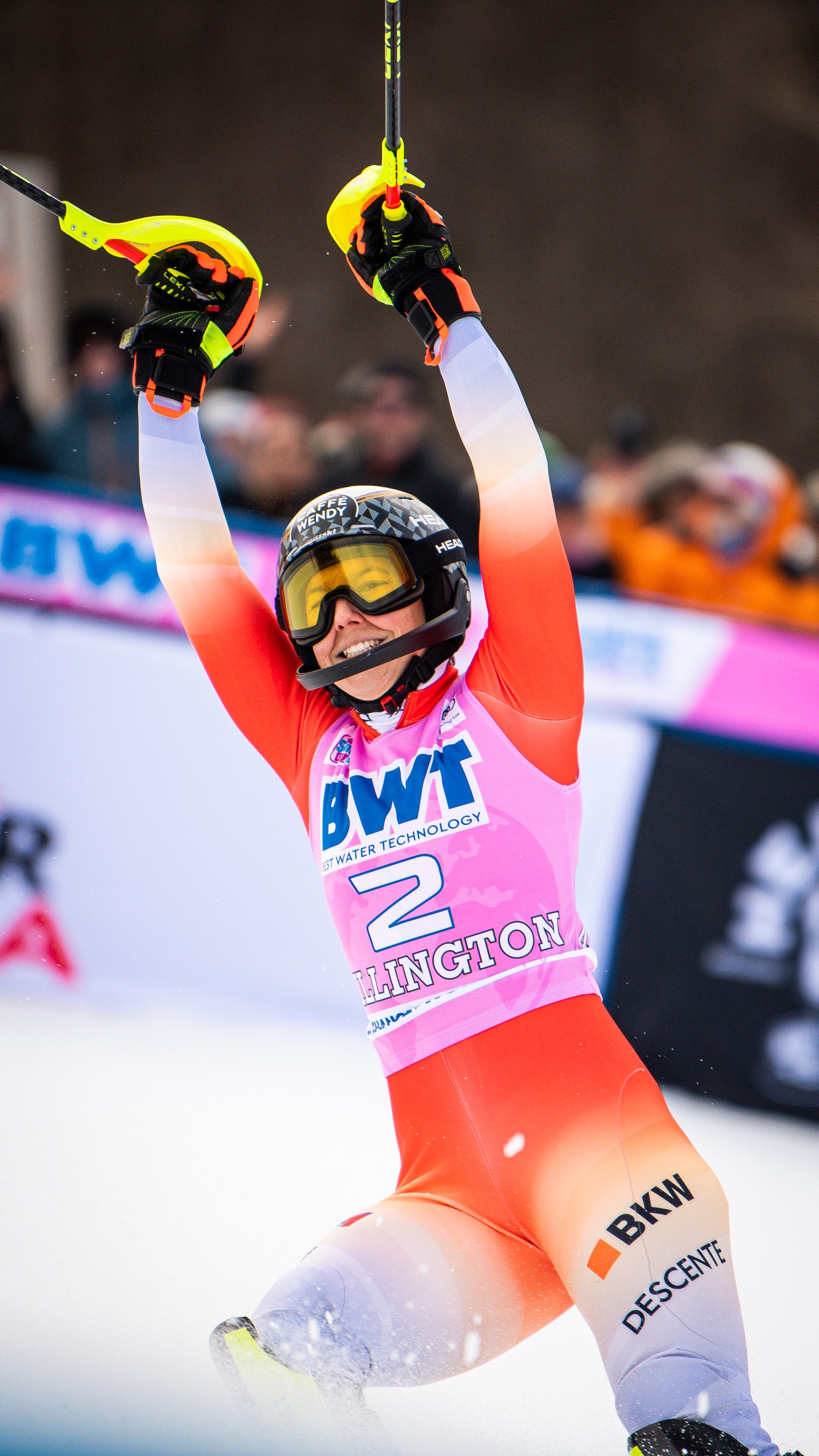 A skier celebrating with arms raised in victory, wearing a black helmet, goggles, and a pink race bib with the number 2, on a snowy course with spectators in the background.