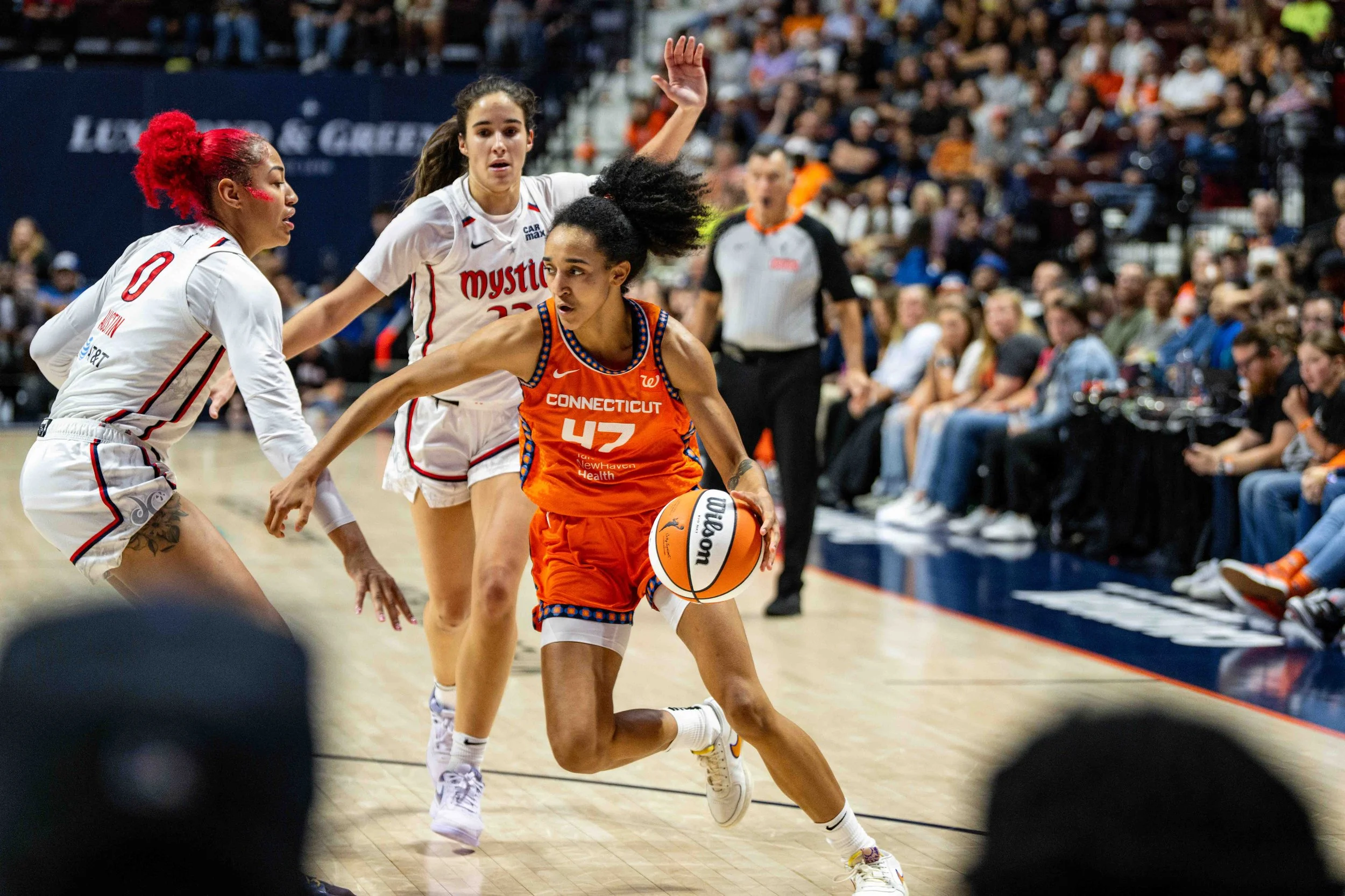 Women’s basketball game, player from Connecticut in orange uniform dribbling basketball, two players from opposing team in white uniforms defending, crowd watching in the background.