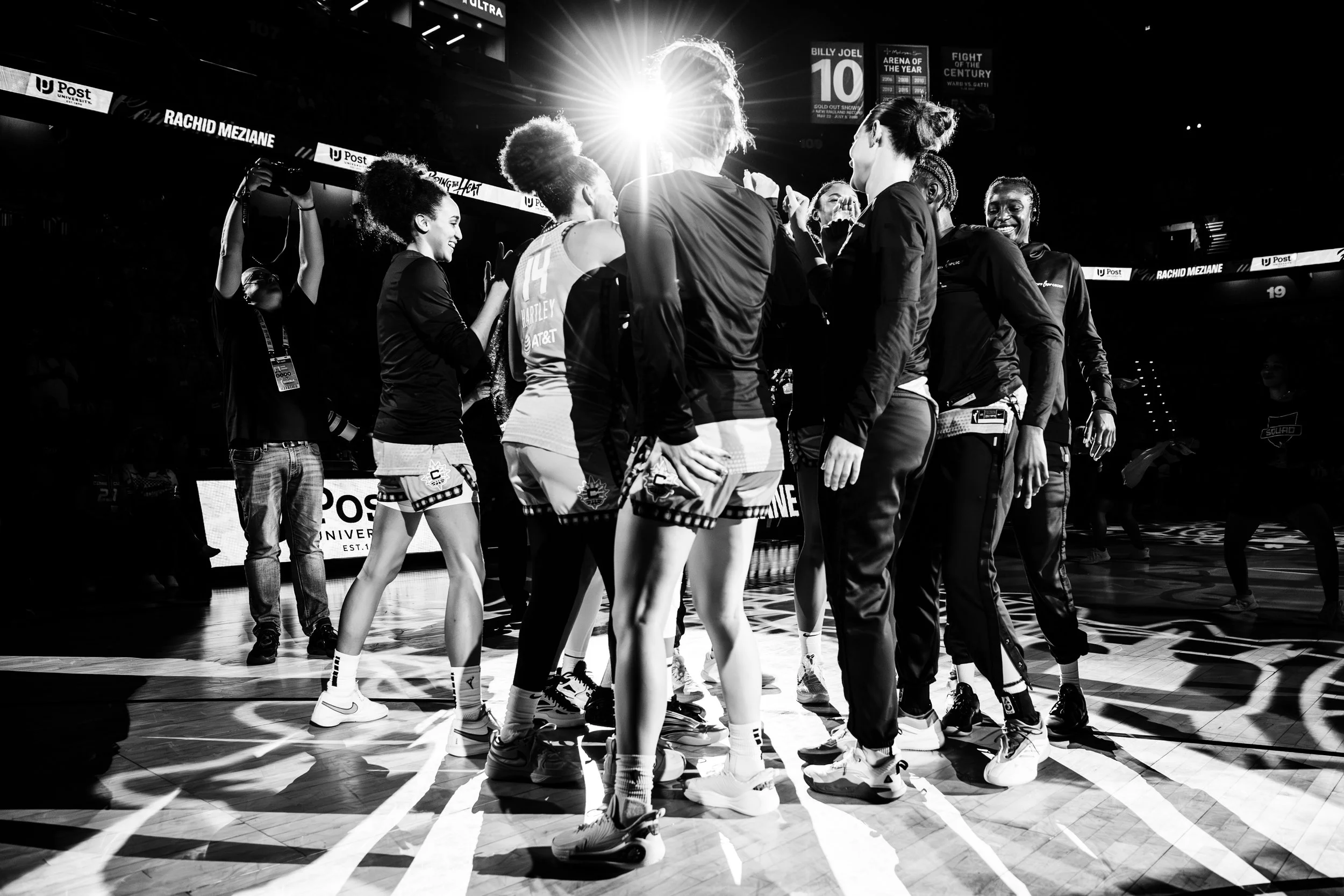 A group of female basketball players on the court, celebrating during a game, with bright light shining behind them.