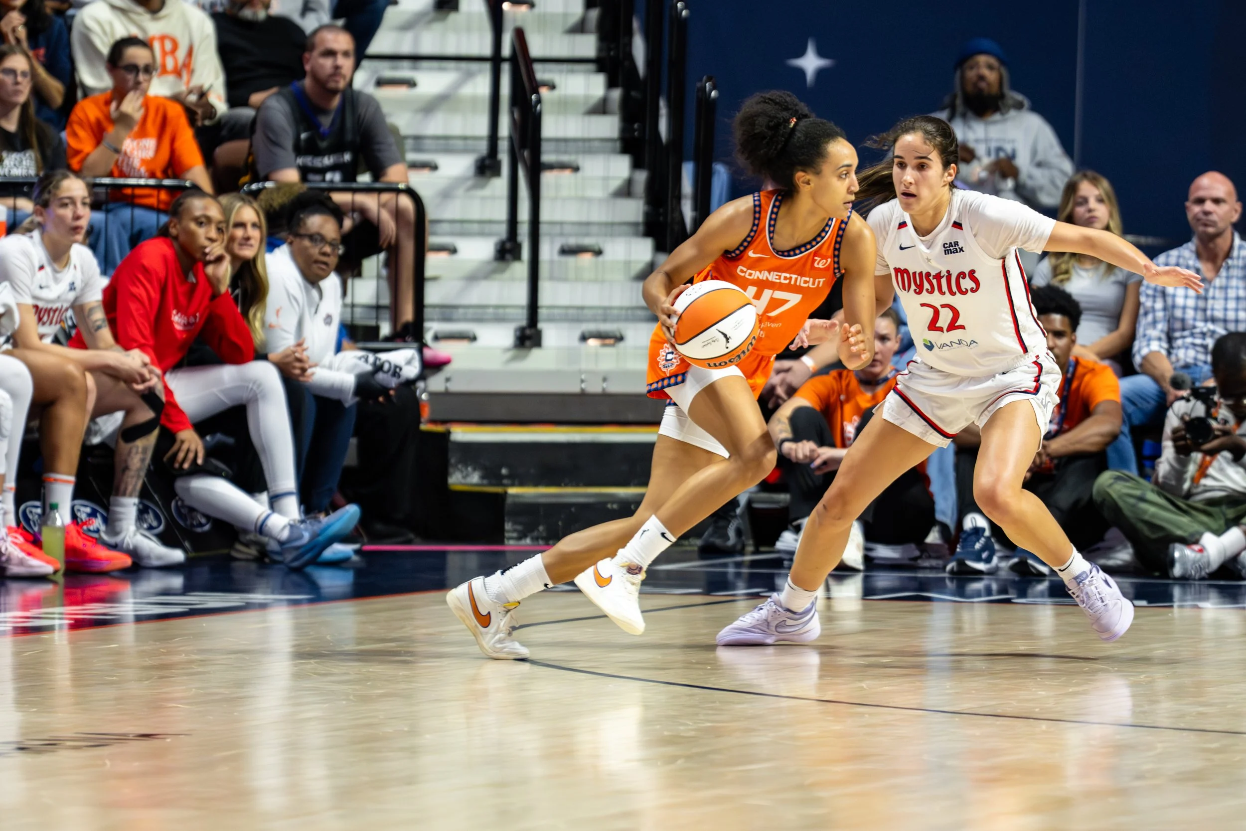 Two female basketball players competing for possession of the ball during a game, with spectators watching from the sidelines.