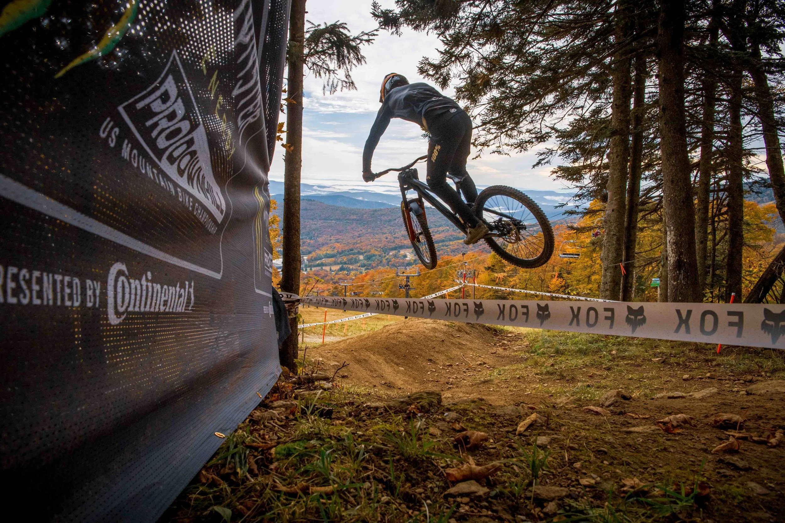 A mountain biker in mid-air jumps over a dirt trail surrounded by trees during a downhill mountain biking race, with autumn foliage in the background.