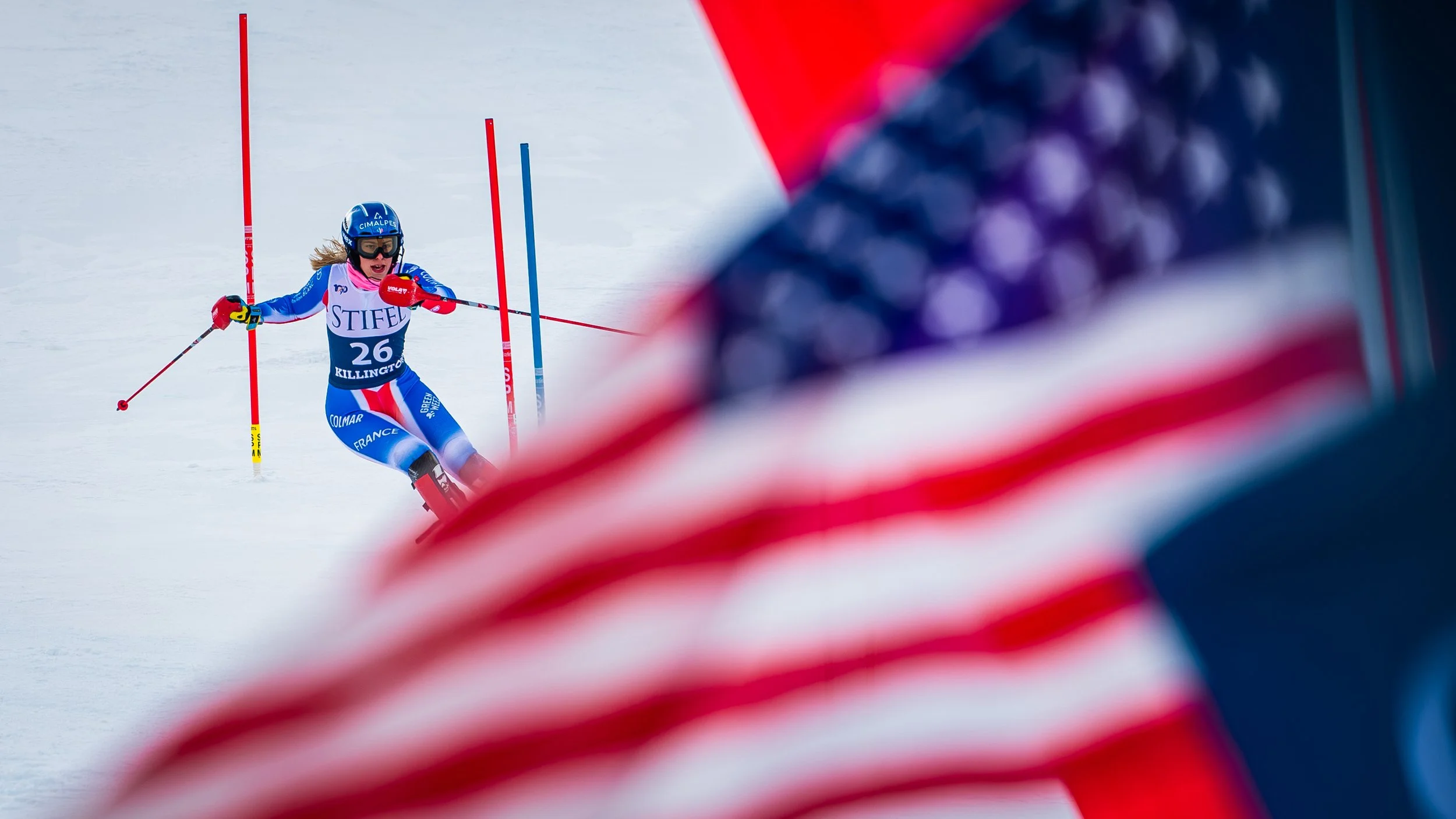 A female alpine skier in a blue uniform with the number 26, racing down a snowy course, navigating between red and blue slalom poles, with an American flag in the foreground.