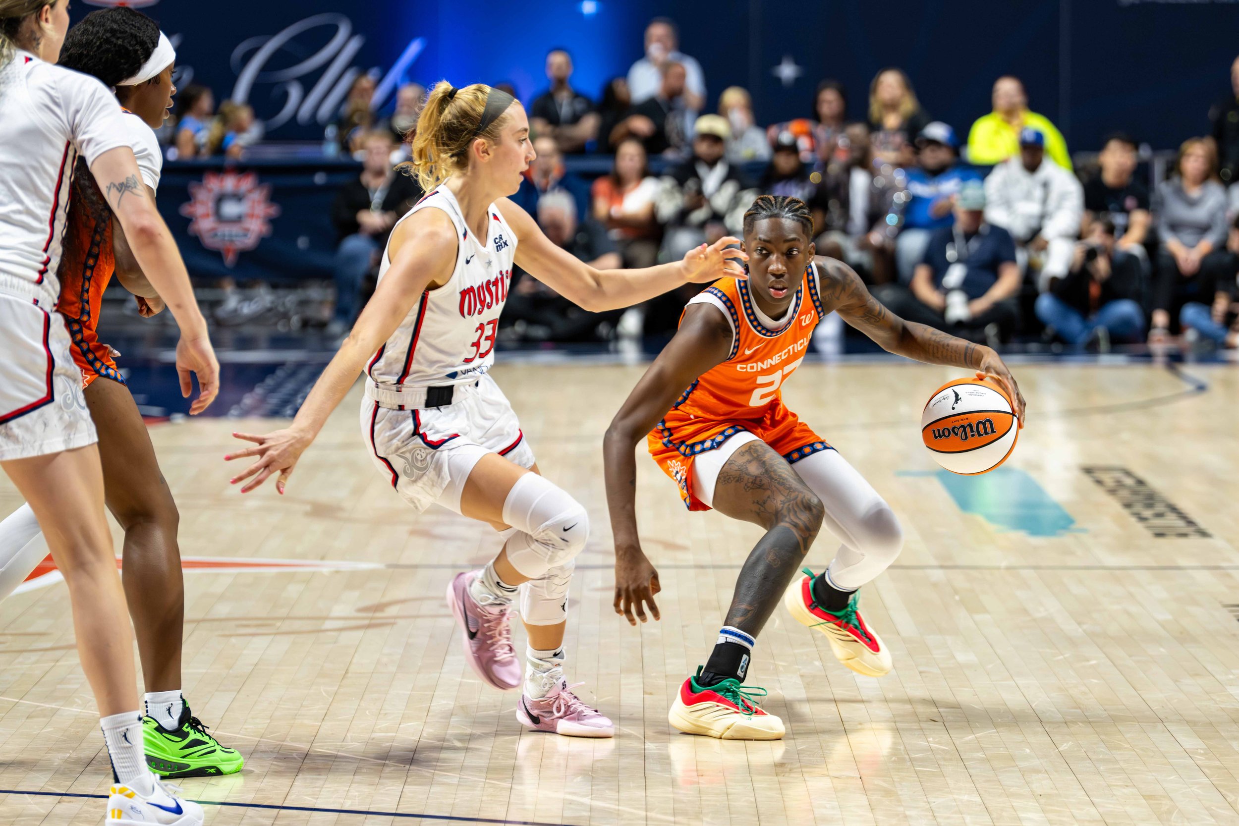 Women playing basketball on court during game with spectators in background.