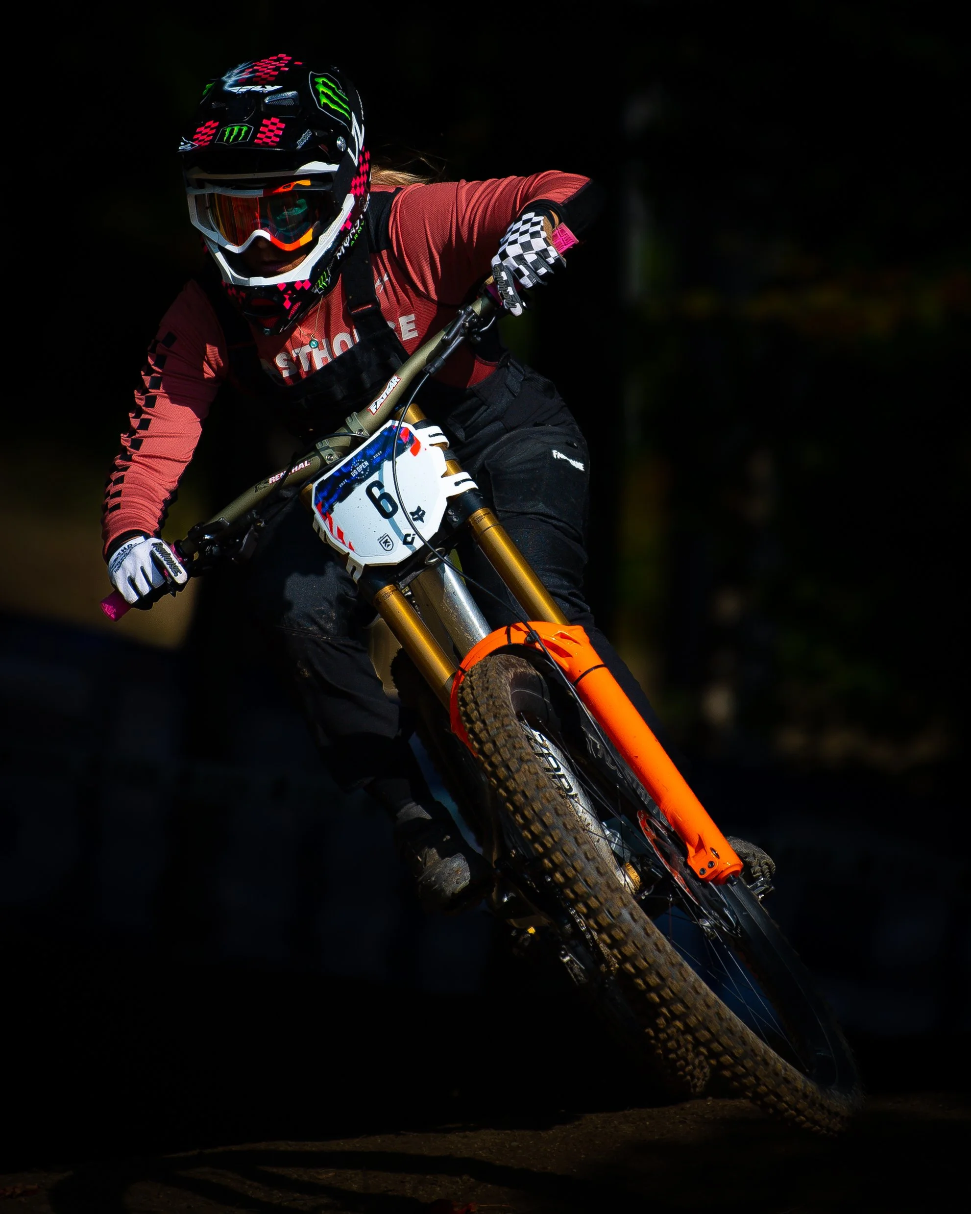 A mountain biker wearing a black helmet with pink and green logos, goggles, a red jersey, black gloves, black pants, and riding an orange mountain bike on a dark trail.