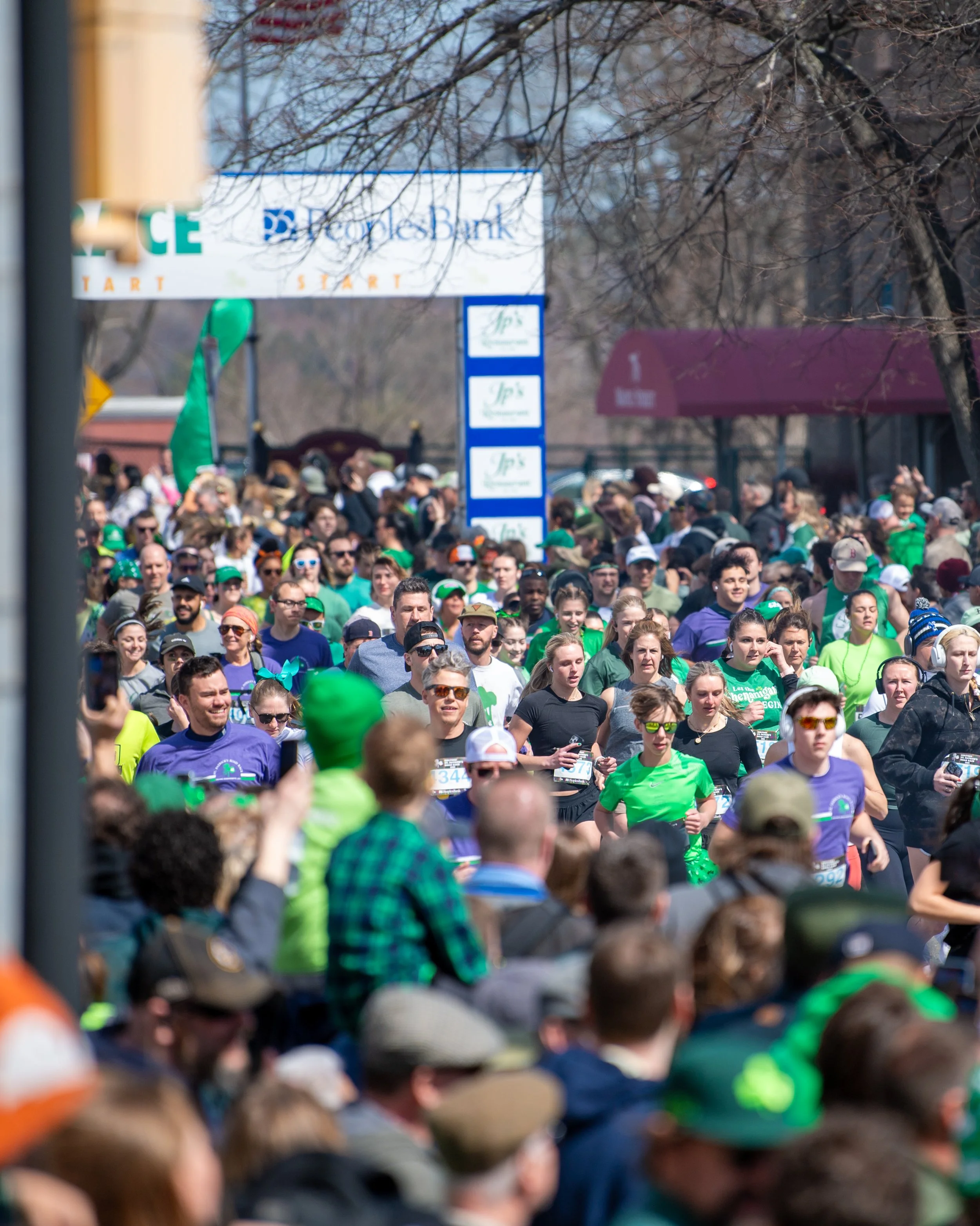 Crowd of marathon runners participating in a race outdoors on a sunny day, some wearing green and purple shirts and sunglasses, with spectators watching on the sidelines.