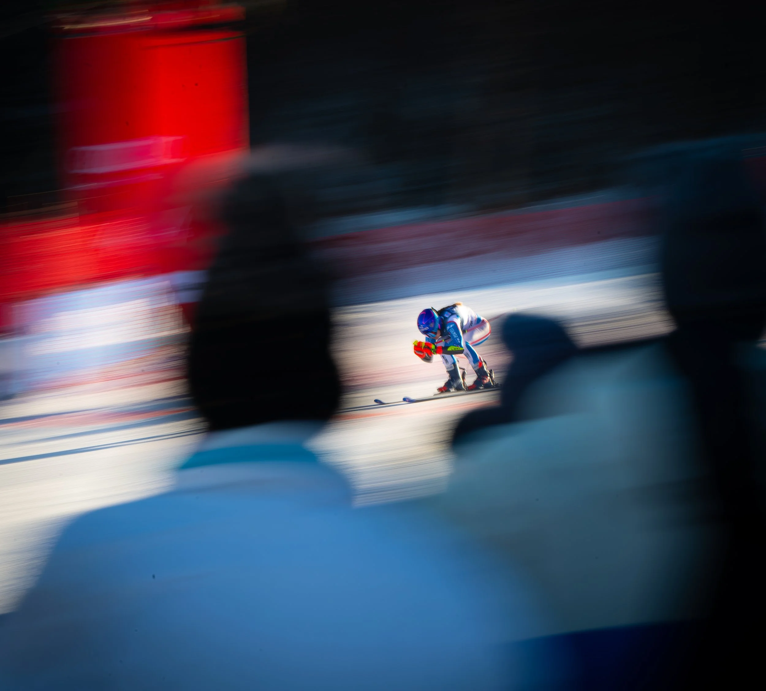 A skier in racing gear skiing on snow, seen through blurred silhouettes of spectators in the foreground.