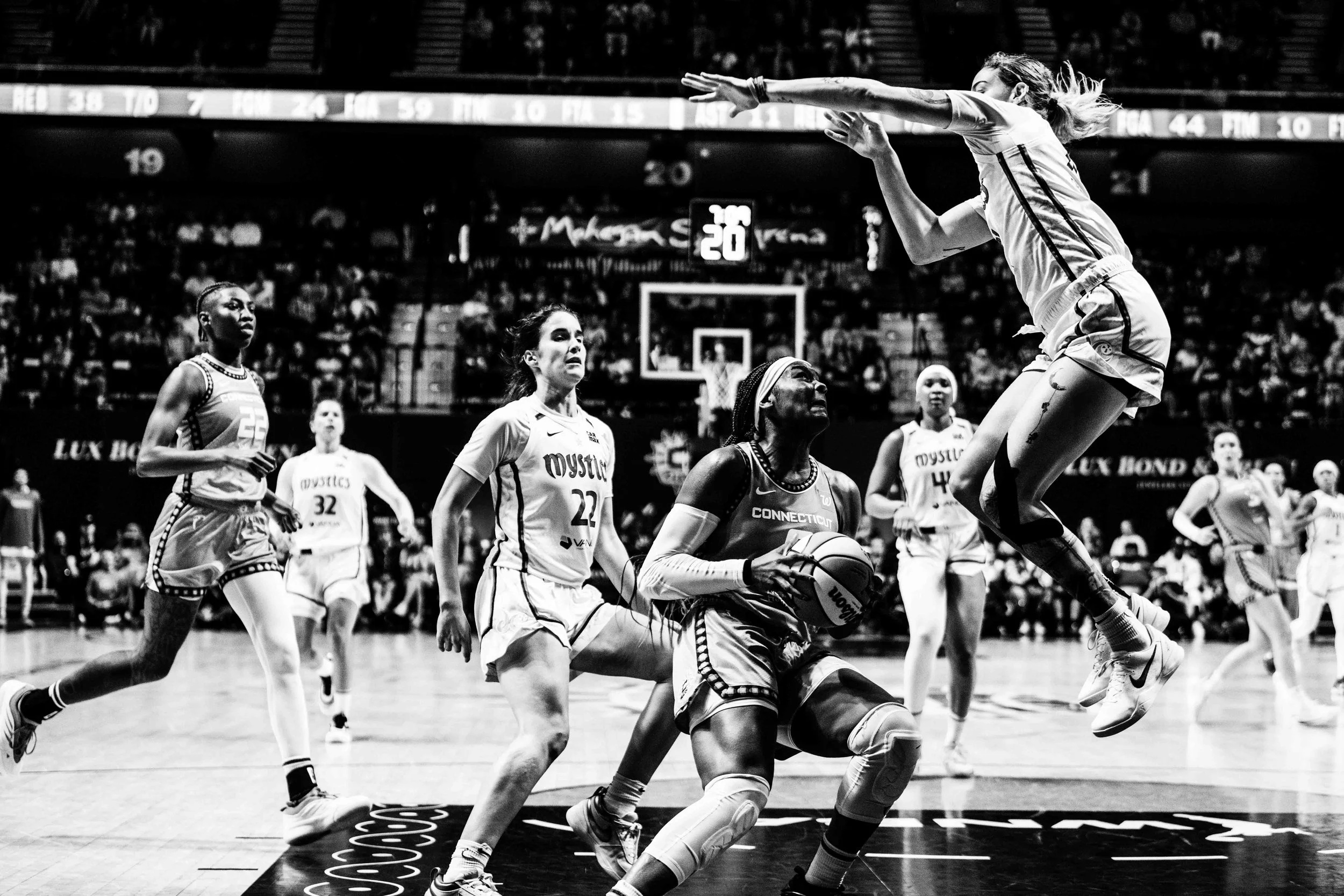 Women playing basketball on the court during a game, with one player jumping to shoot or block, and others observing or preparing to rebound, in an indoor arena.