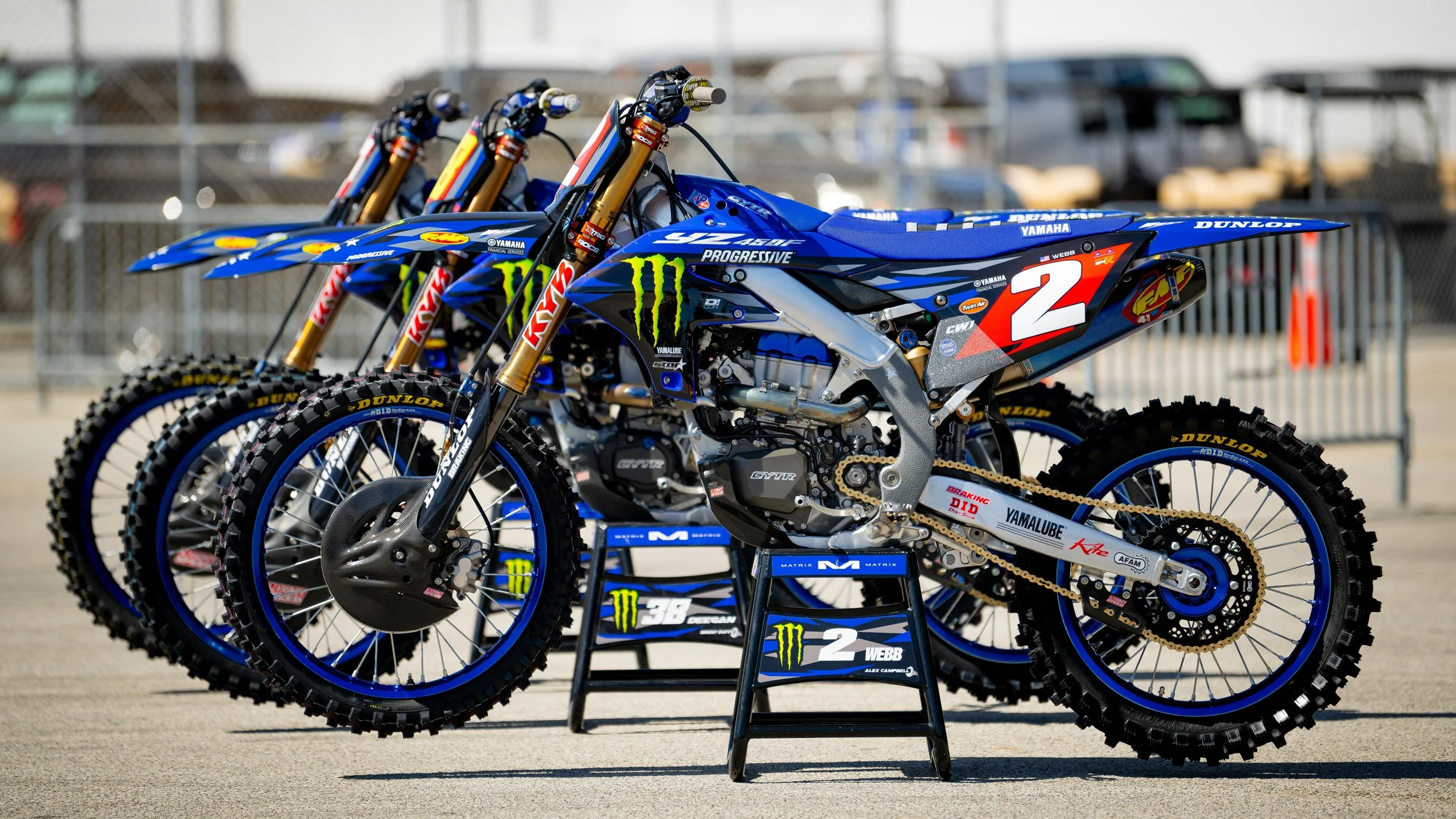 Three blue Yamaha dirt bikes with number 2 decals, positioned on a stand, in a paddock area with fences and parked vehicles in the background.