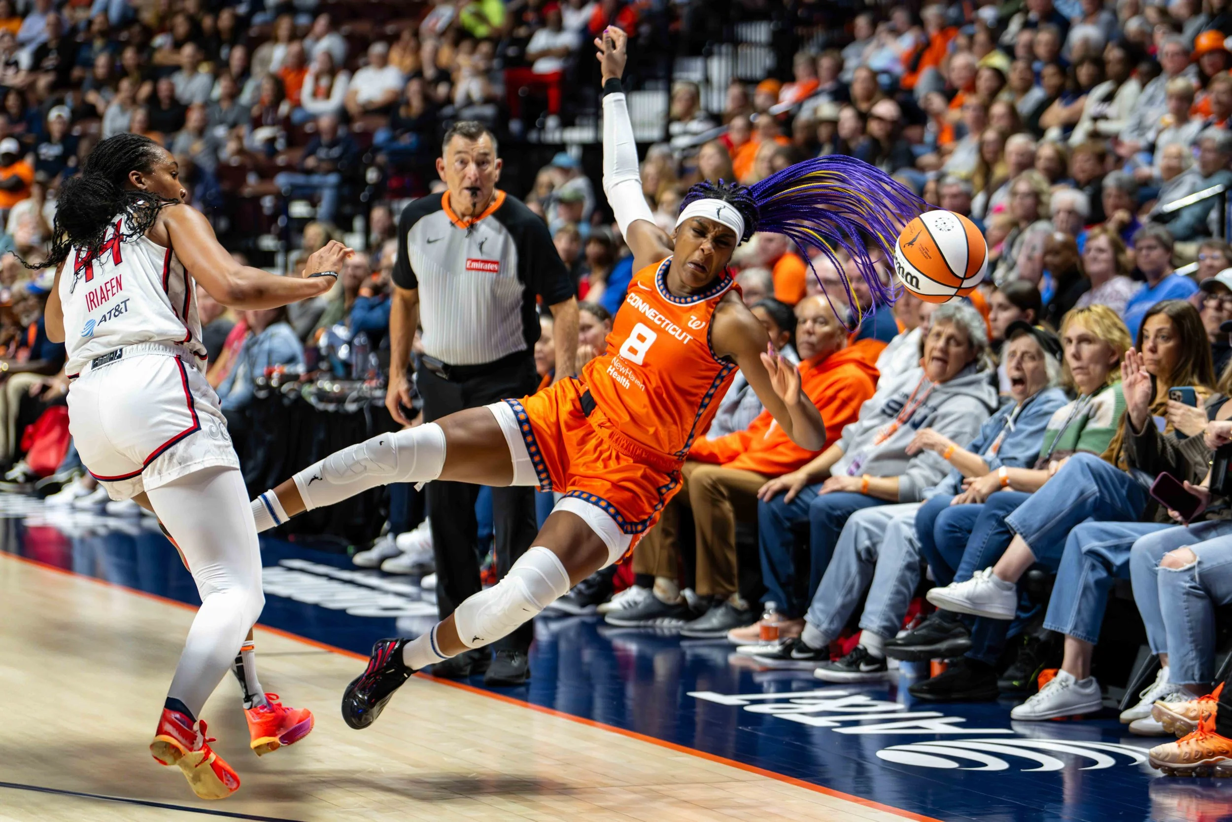 A female basketball player in an orange uniform, number 9, is falling to the floor while reaching for a basketball near the sideline during a game. A player in a white uniform is nearby, with spectators watching from the stands.