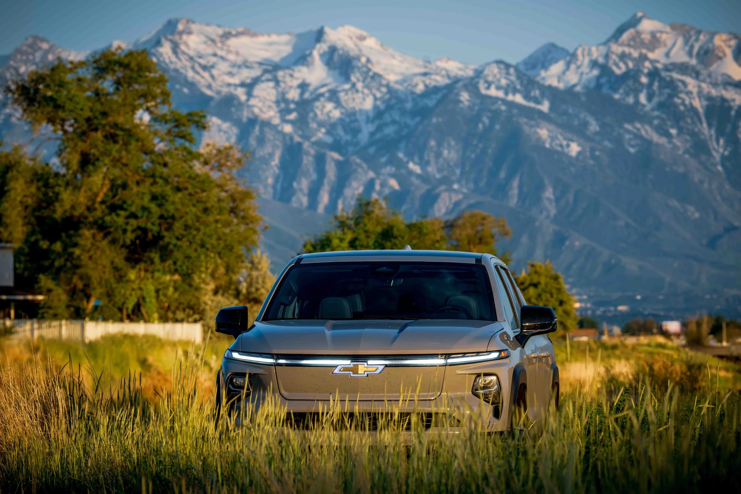 A silver Chevrolet SUV parked in a grassy field with trees and snow-capped mountains in the background during sunset.