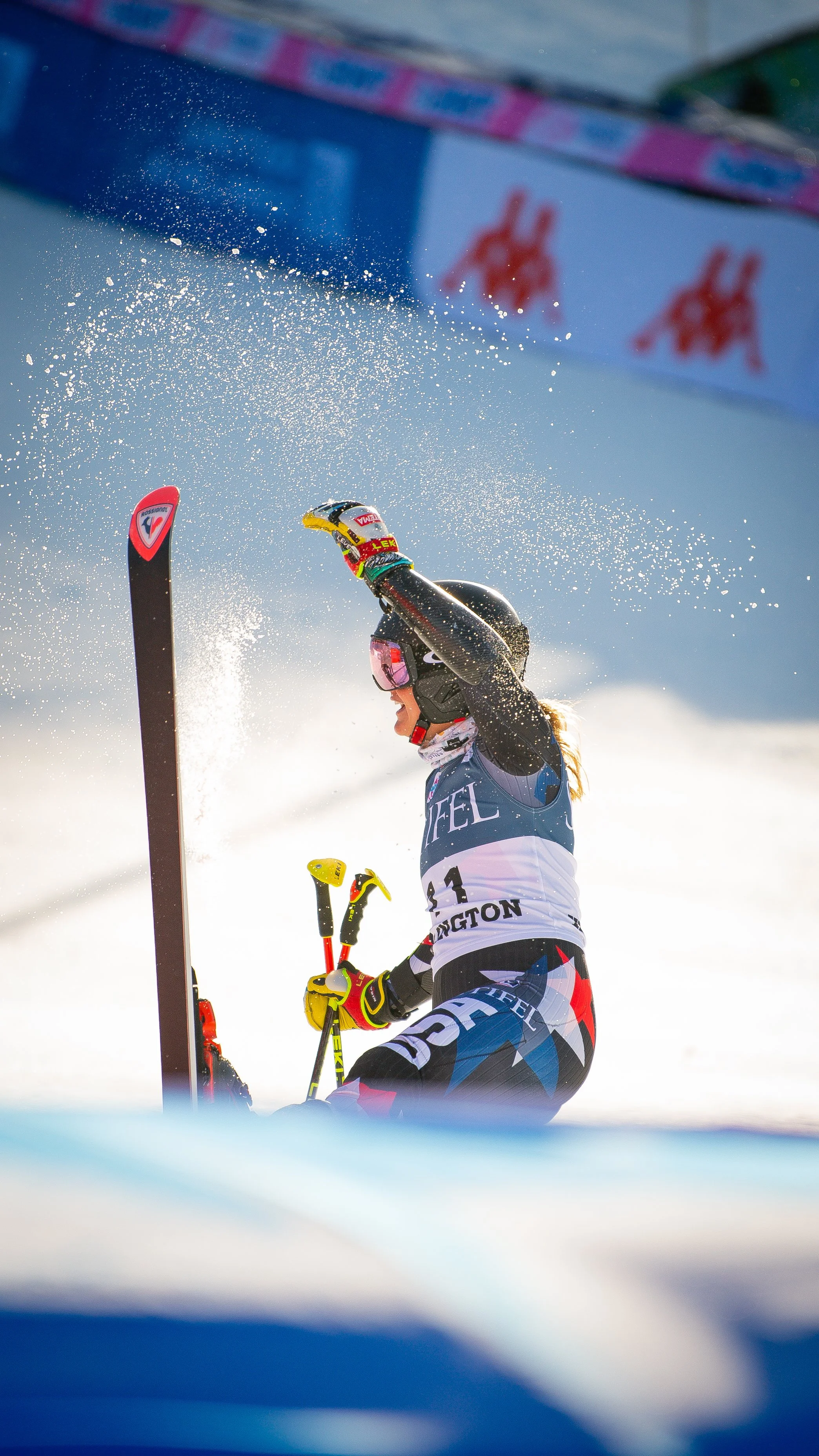 A female skier kneeling in the snow, celebrating after a race, with snow spraying around her and a blurred background of advertising banners.