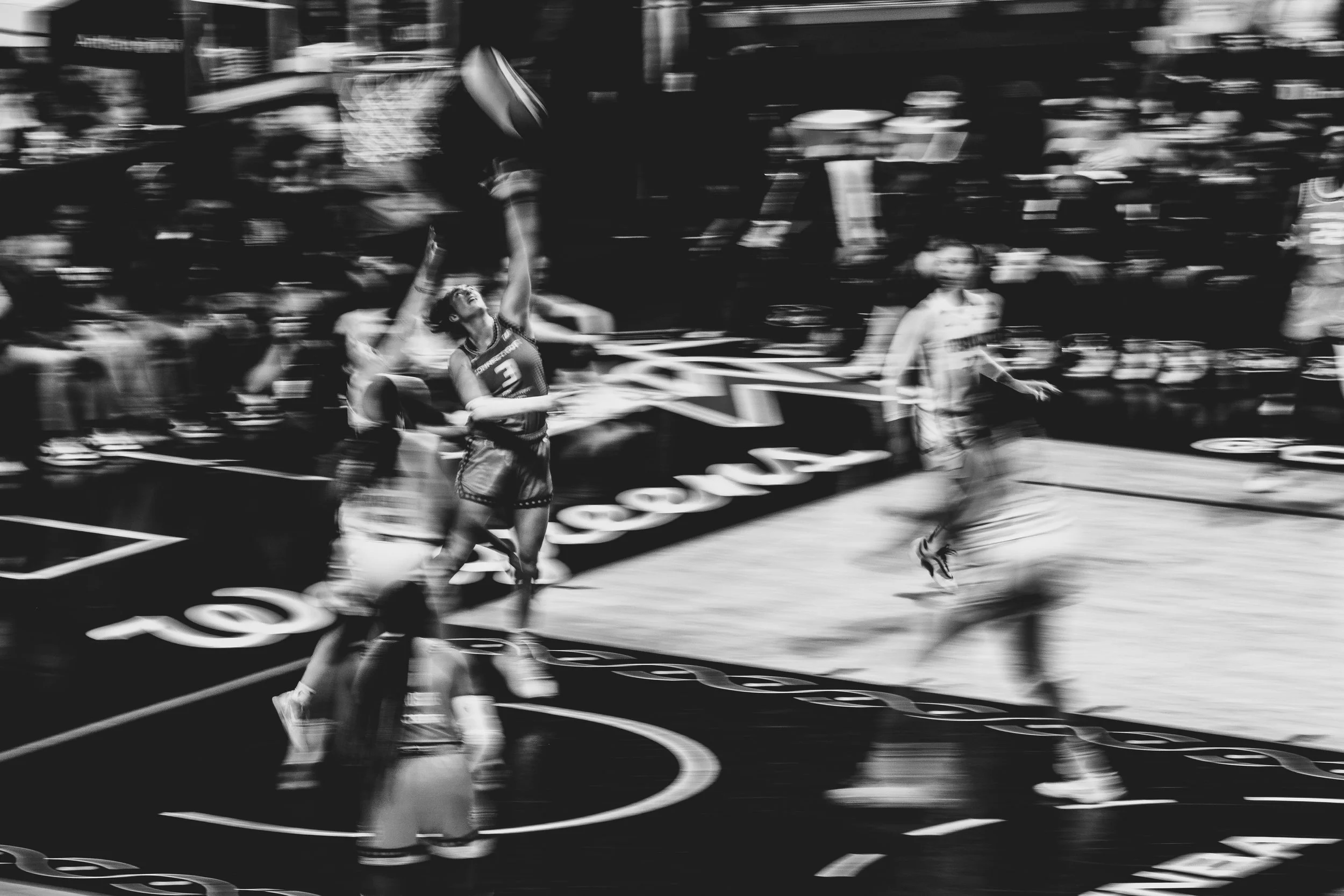 Black and white action shot of a women's basketball game with a player jumping to shoot the ball near the basket, other players around her and spectators in the background.
