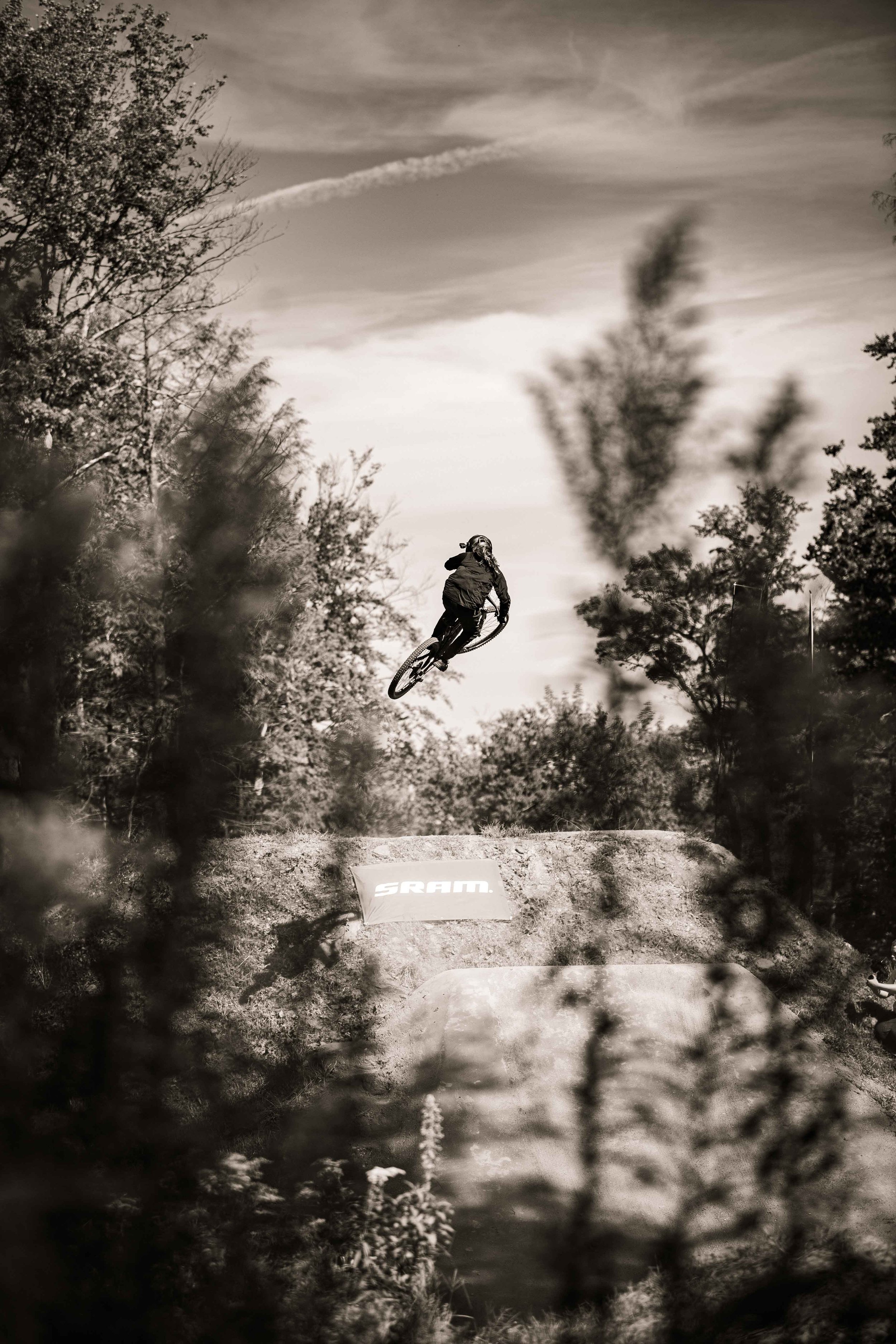 A person riding a mountain bike in mid-air, performing a trick on a dirt jump trail surrounded by trees, with a partly cloudy sky overhead.