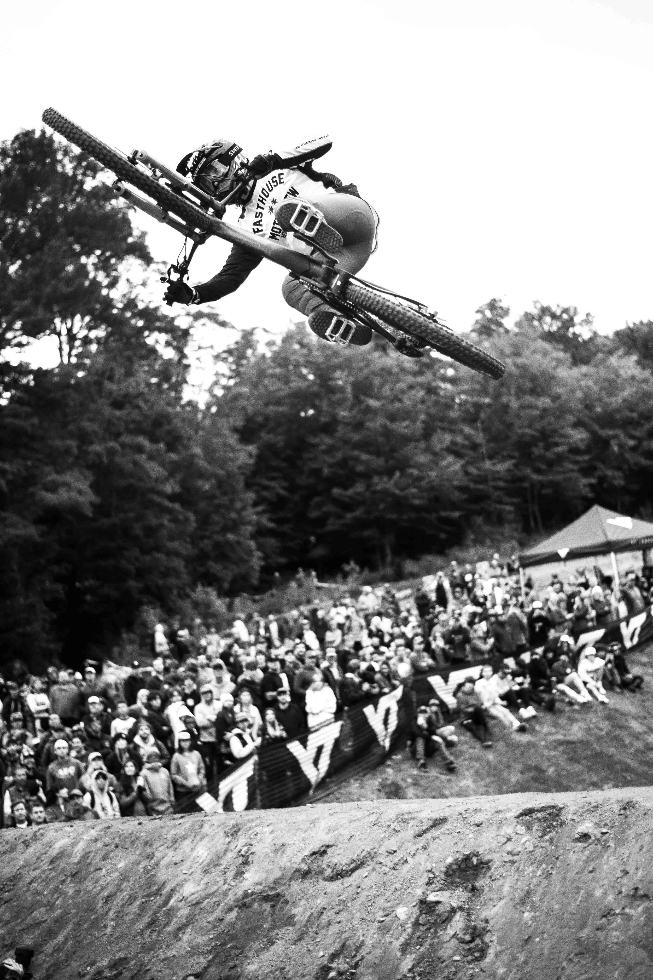 A mountain biker in mid-air performing a stunt over a dirt trail during a race, with a crowd watching from behind a barrier and trees in the background.