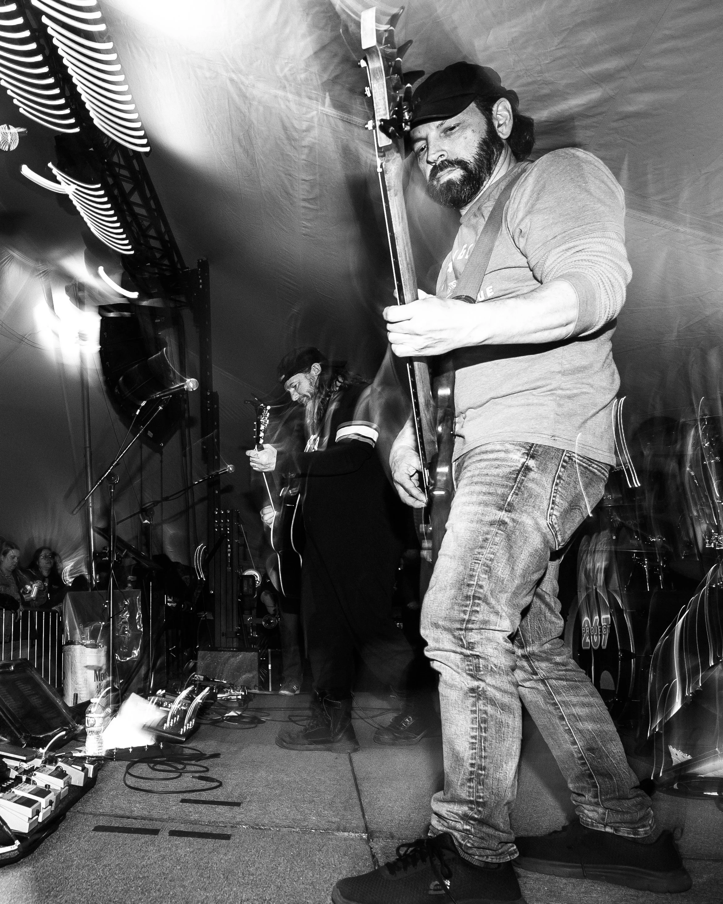 Black and white photo of two musicians performing on stage. The musician in the foreground is playing an electric guitar, wearing a beret and casual clothes. The musician in the background is playing an acoustic guitar, with long hair and a hat, and 