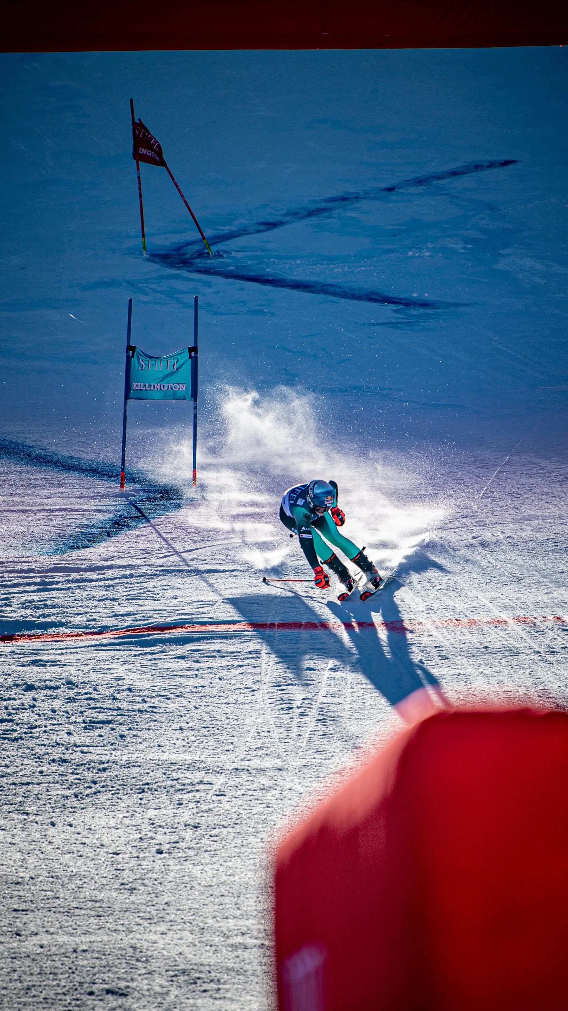 A skier wearing a helmet and teal ski suit navigates through a slalom course marked by blue and red gates on a snowy slope under clear blue skies.