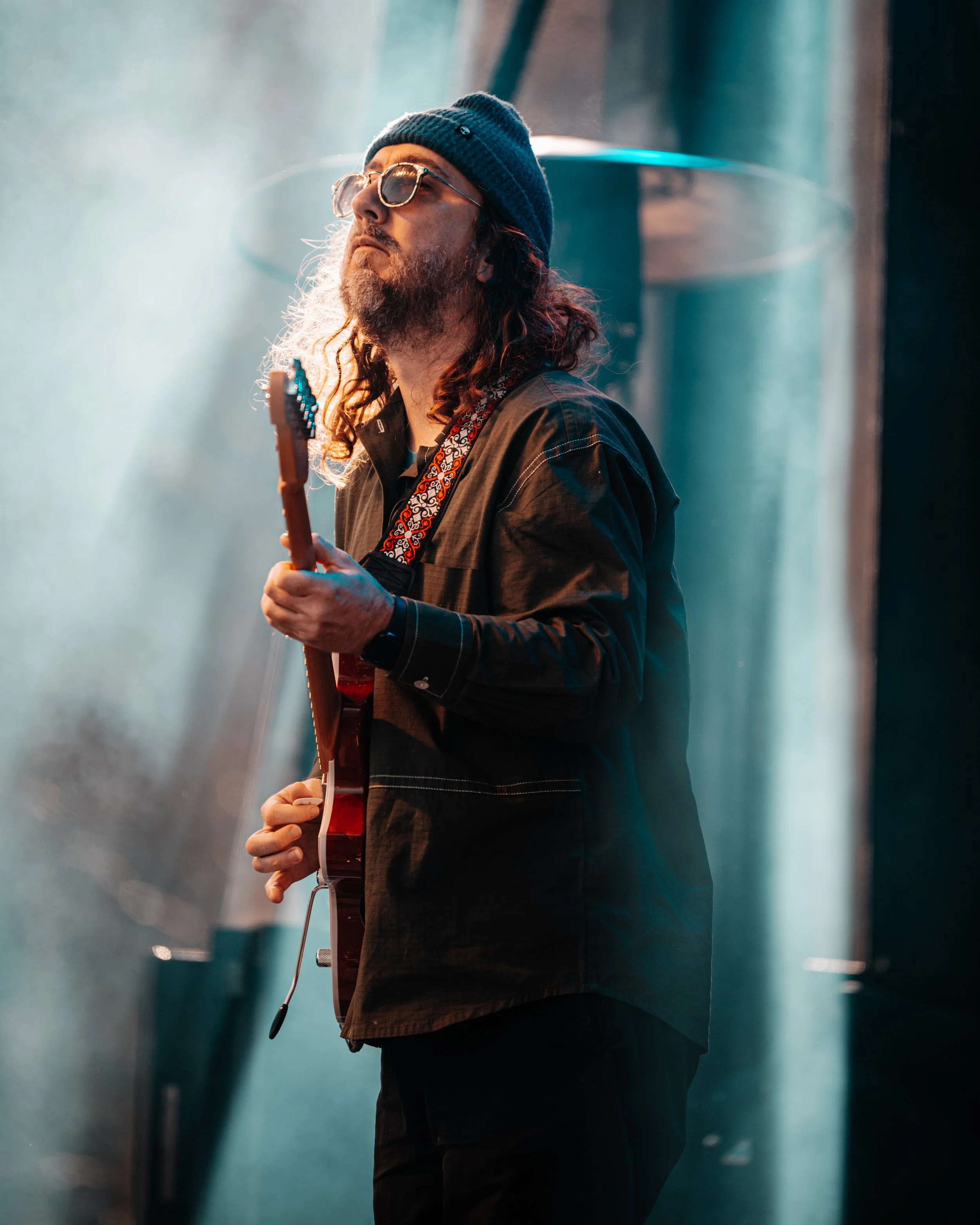 A man with long curly hair, glasses, and a blue beanie playing an electric guitar on stage.