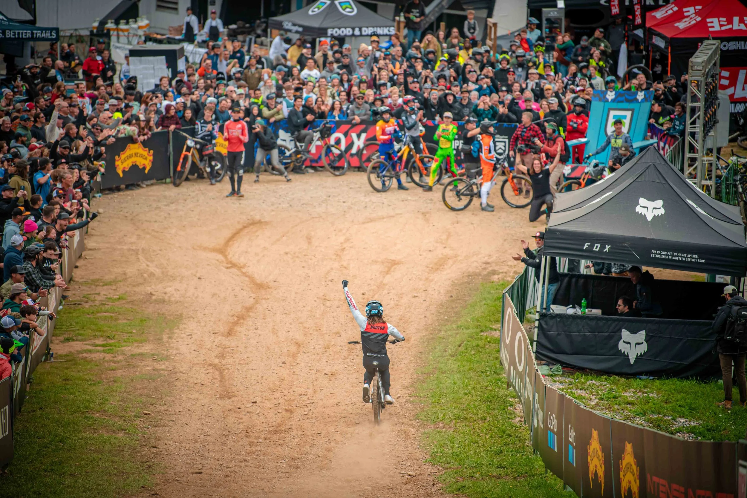 A cyclist in racing gear rides down a dirt track with a crowd of spectators and racers watching from the sides and in the background.