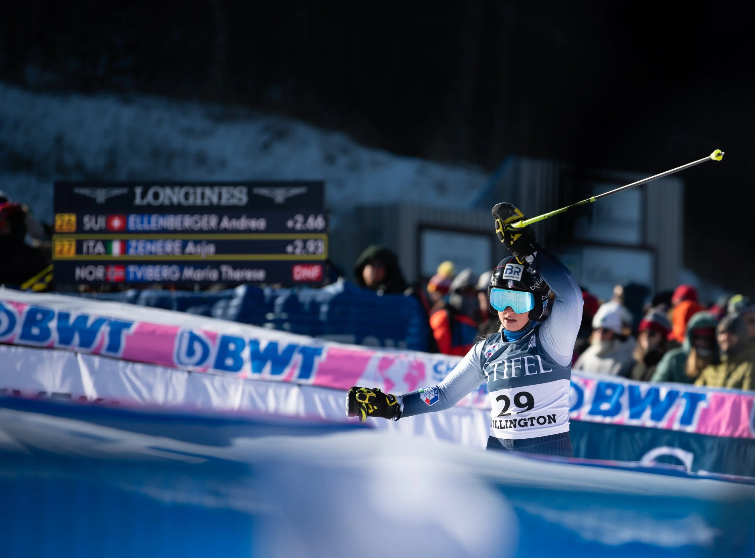 A female alpine skier celebrating after a race, wearing a helmet, goggles, and a race bib with the number 29, raising her ski pole in the air amidst snow and spectators.