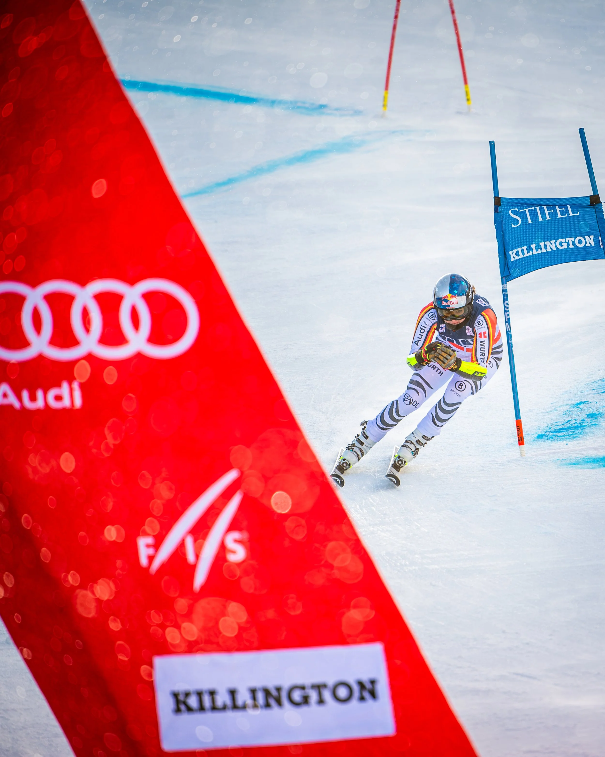 A skier wearing a racing suit and helmet navigates a slalom course on snow, passing a blue gate with 'STIFEL KILLINGTON' on it, in an alpine ski race event.