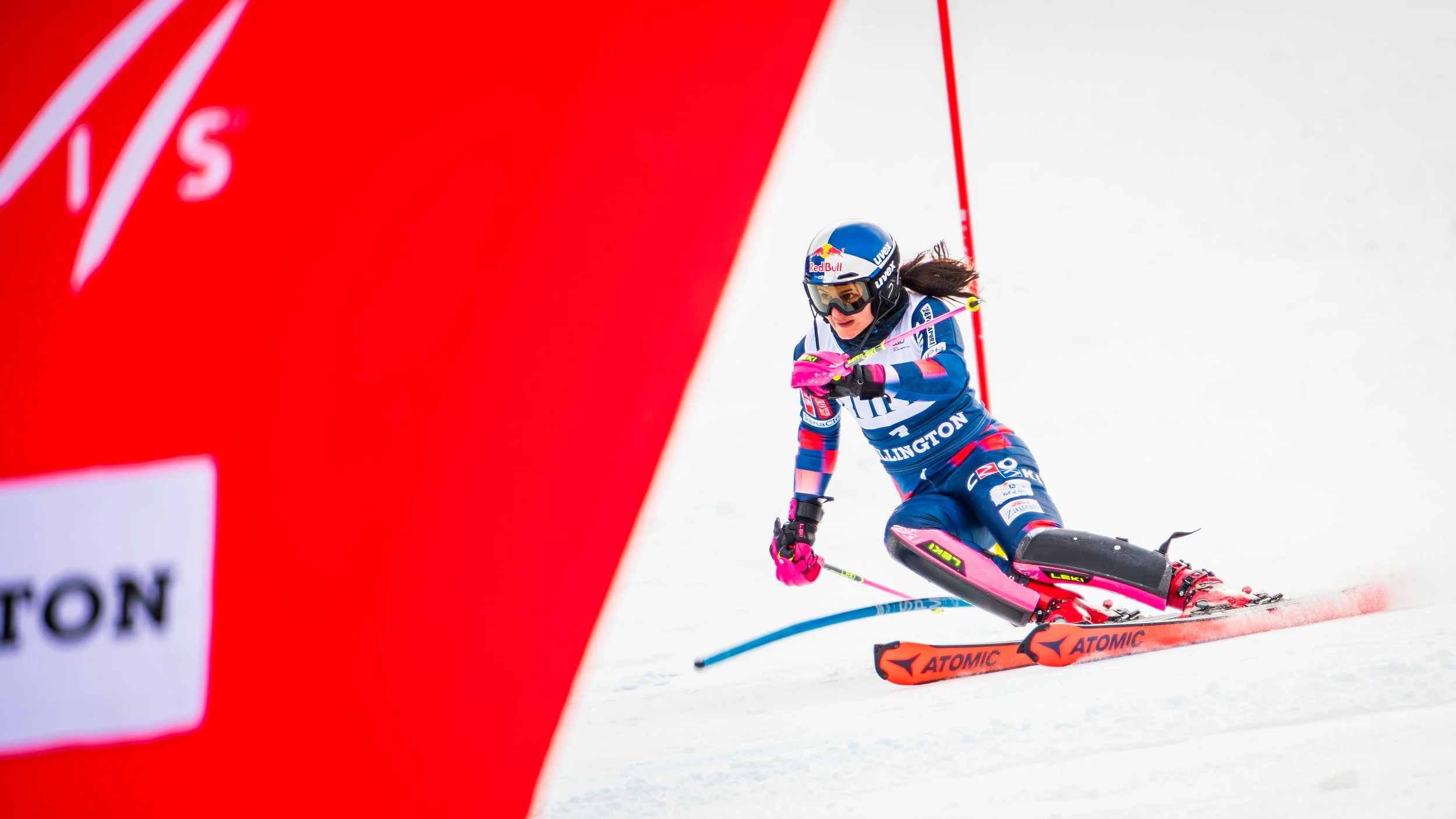 A female alpine skier racing down a snowy slope, wearing a blue racing suit, helmet, and goggles, with a large red wall on the left side and safety netting in the background.