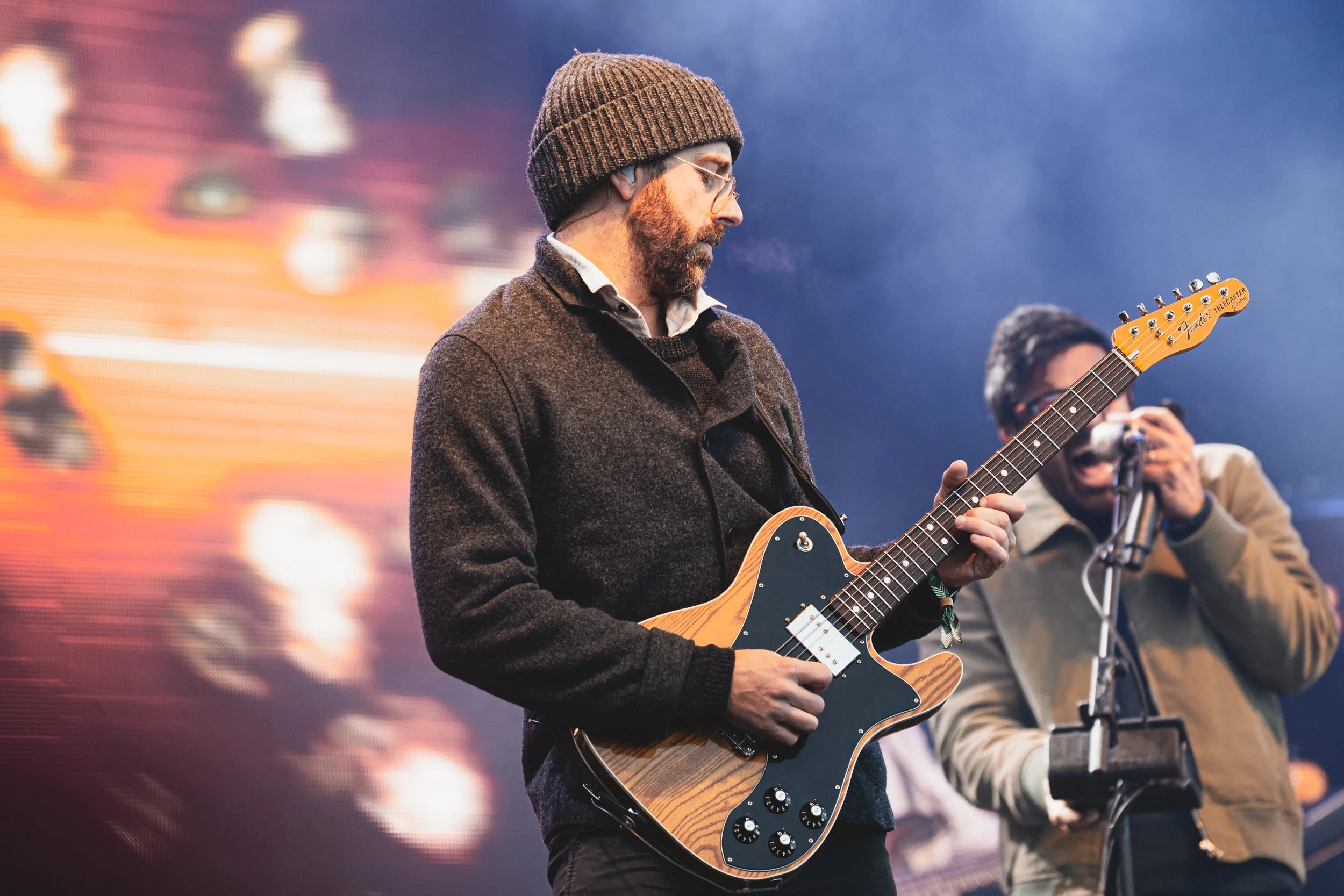 Man wearing glasses, a brown knit cap, and a dark sweater playing an electric guitar on stage with a colorful blurred background.