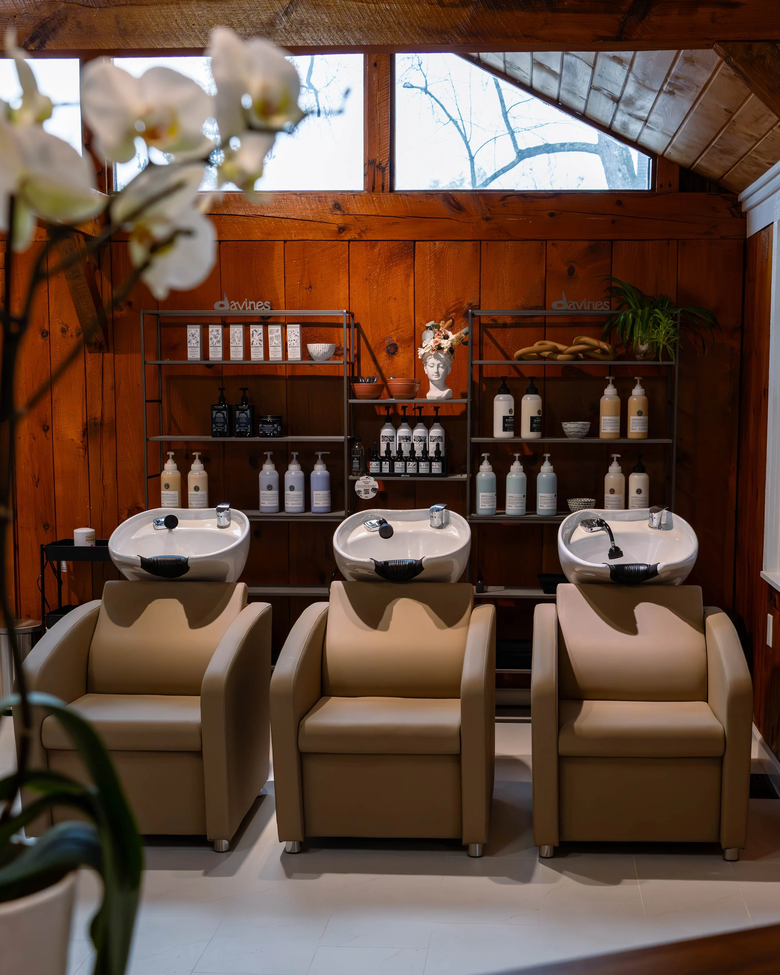Three hair washing stations with beige chairs and white sinks in a salon with wooden wall paneling and shelving containing hair products, decorative items, and plants.
