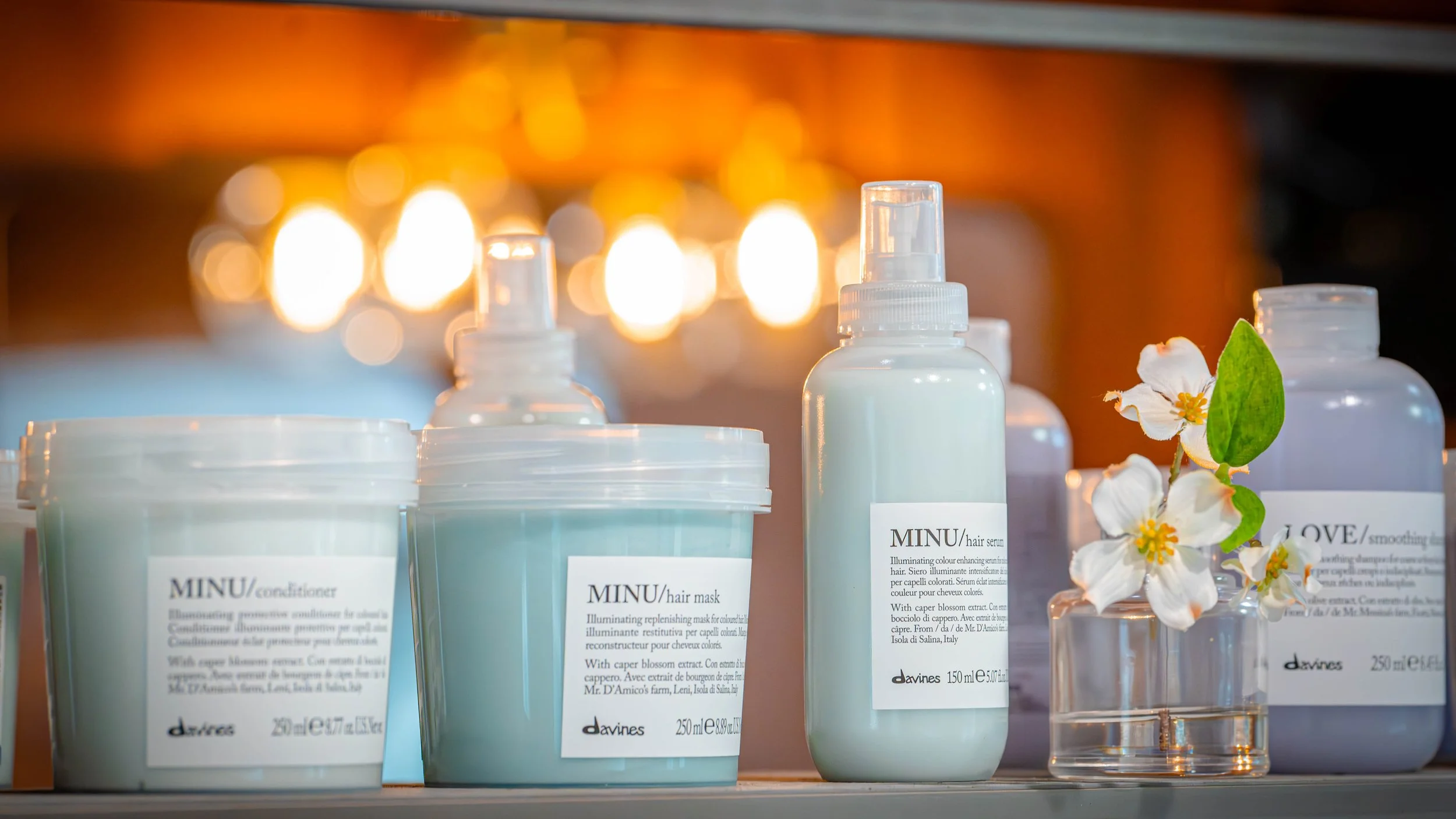 Collection of skincare products including conditioner, hair mask, hair serum, and smoothing cream with a small glass vase of white flowers on a shelf, warm lighting in the background.